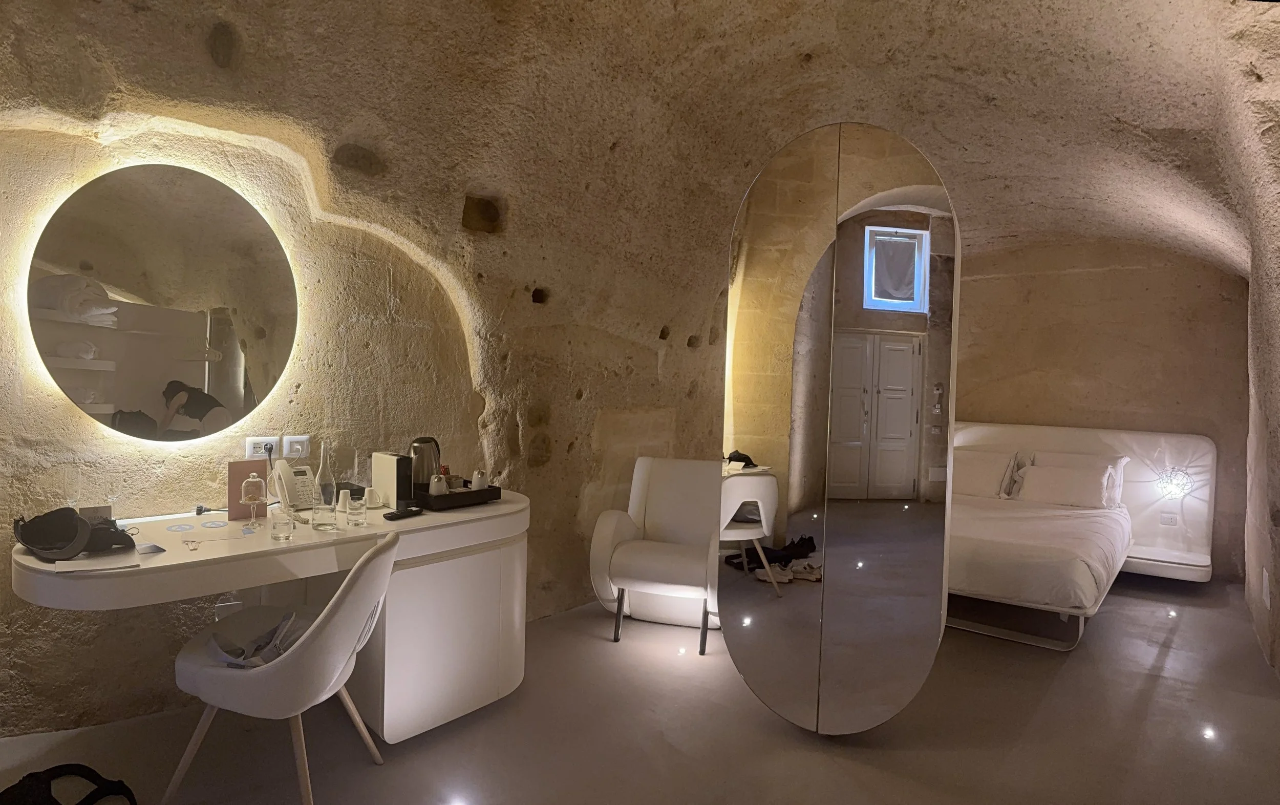 Interior of a room with textured stone walls, a glowing circular mirror, a white table with bottles and glasses, a white chair, a mirror, and a bed with white bedding.