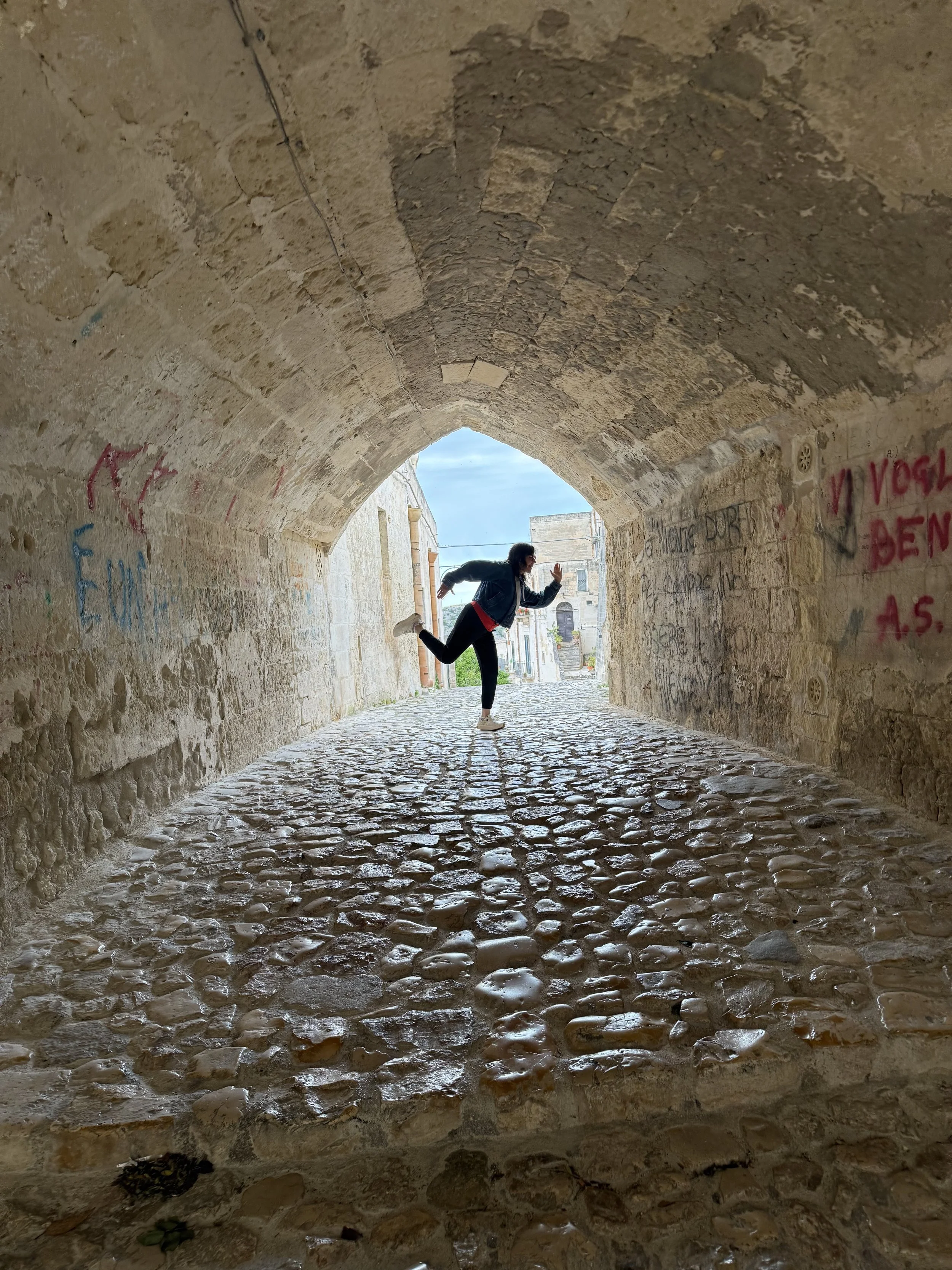 A person running and jumping in an arched stone tunnel with graffiti on the walls and a cobblestone floor.