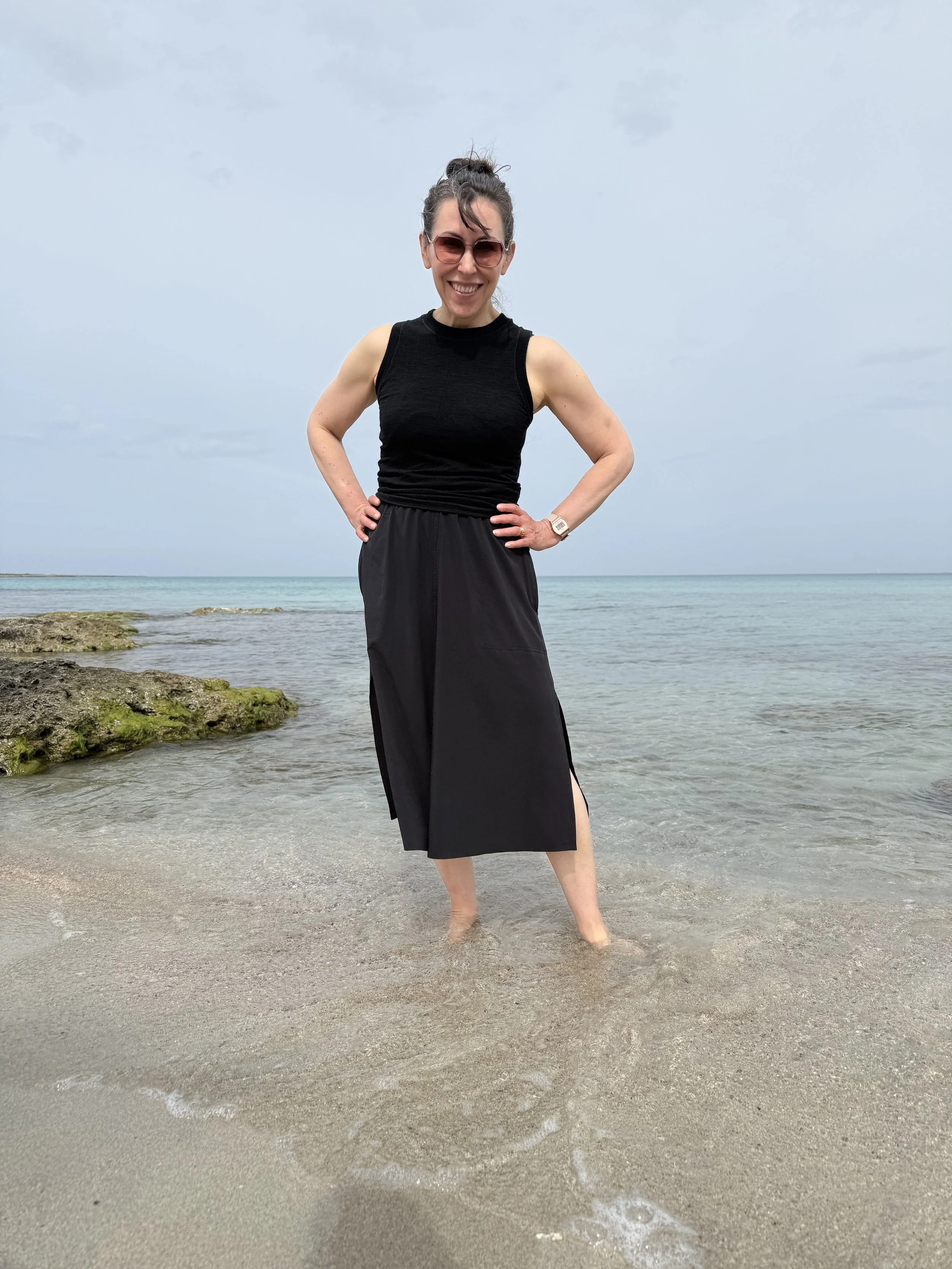 A woman standing in shallow water at the beach, smiling, wearing sunglasses, a sleeveless black top, and black skirt, with rocks and the ocean in the background.