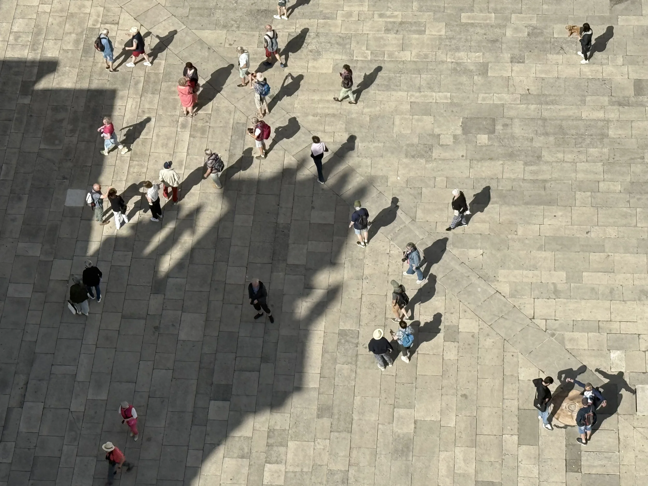 An aerial view of a large stone-paved outdoor area with scattered people walking and standing, casting shadows.