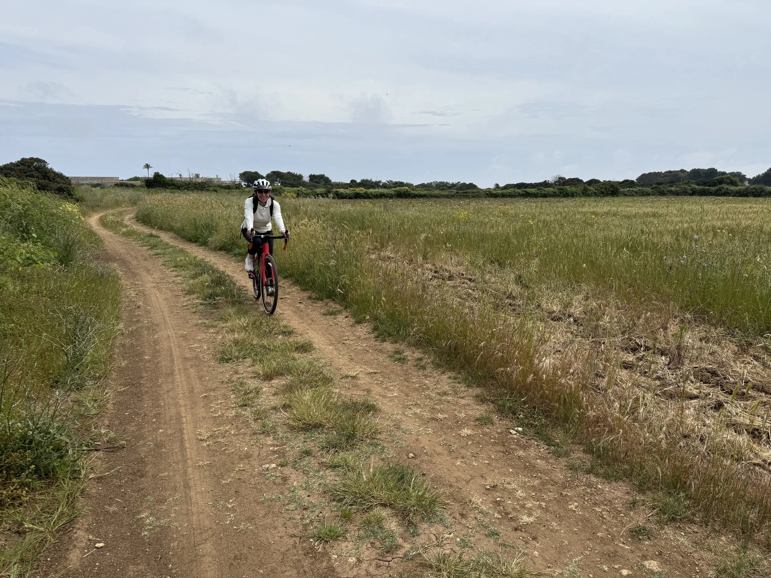 A woman riding a bicycle along a dirt trail through a grassy field on a cloudy day.
