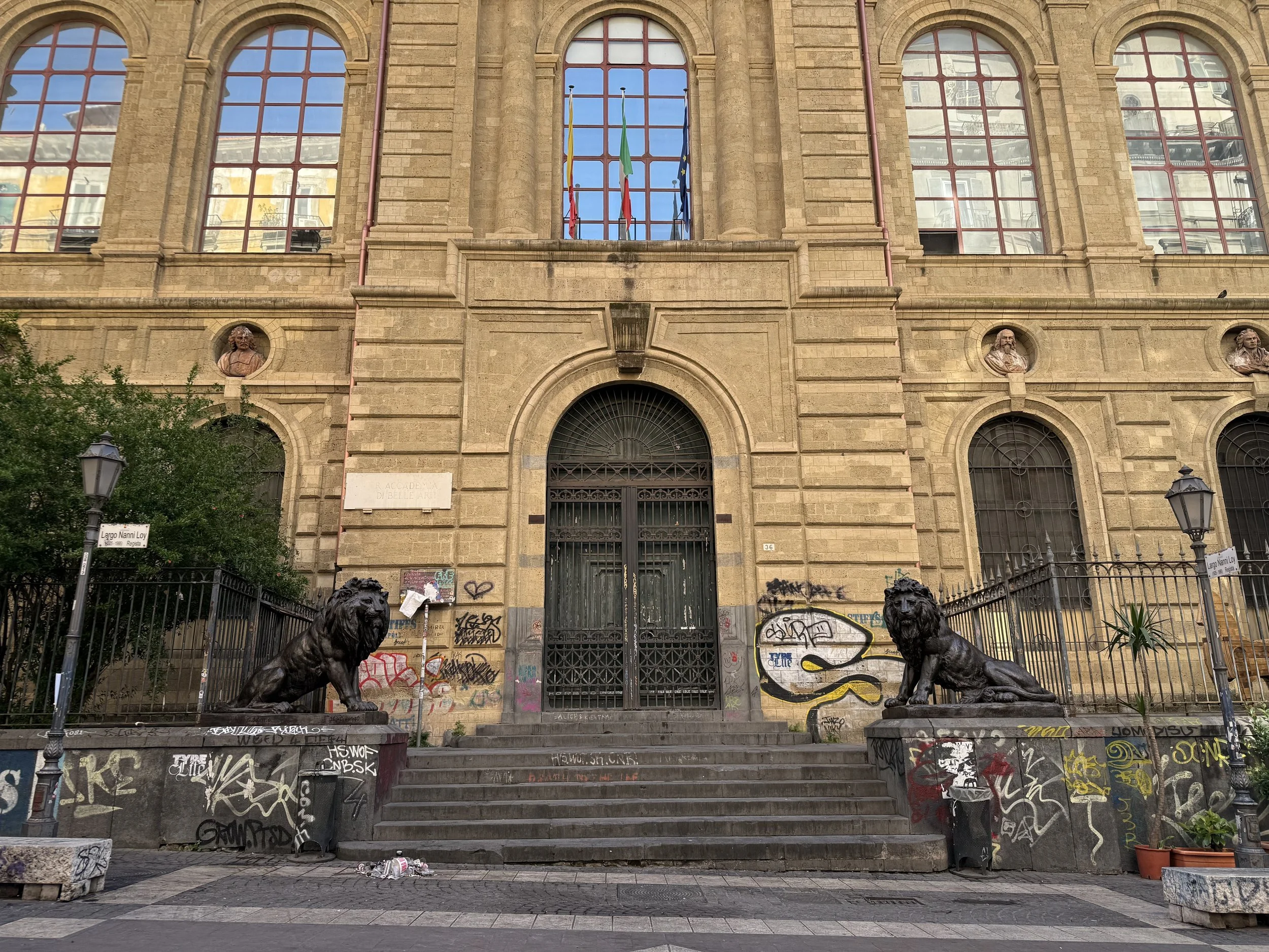 A historic building with large arched windows, adorned with three bust sculptures above the entrance. In front, there are two statues of lions on pedestals, a set of stairs, graffiti on the lower part, and street signs indicating "Largo Nanni Loy" an