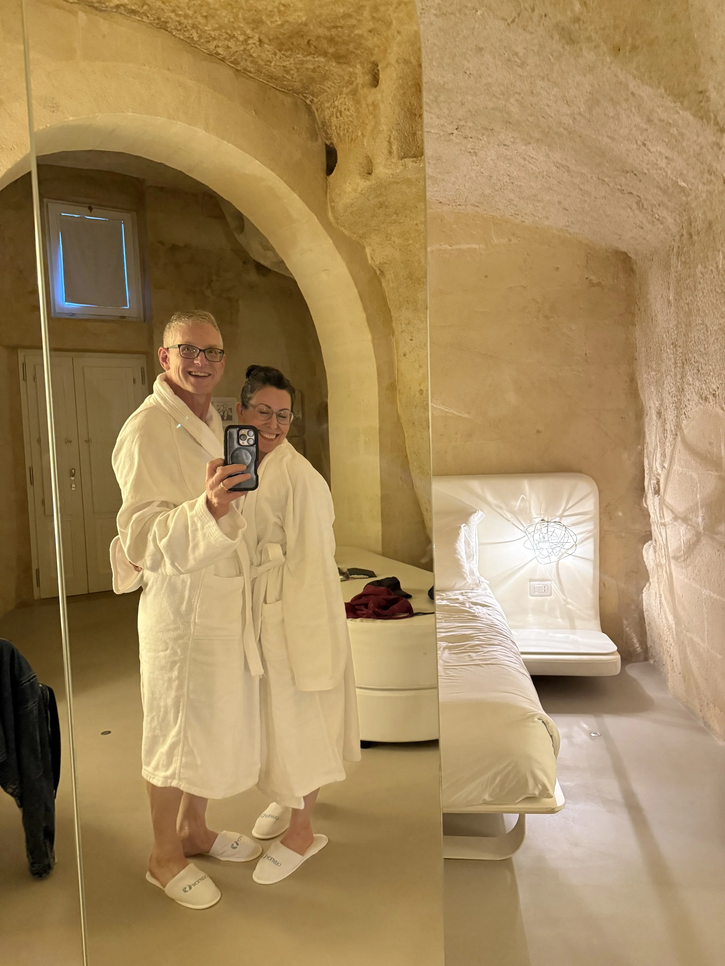 A man and a woman in white bathrobes taking a mirror selfie in a room with stone walls and a bed.