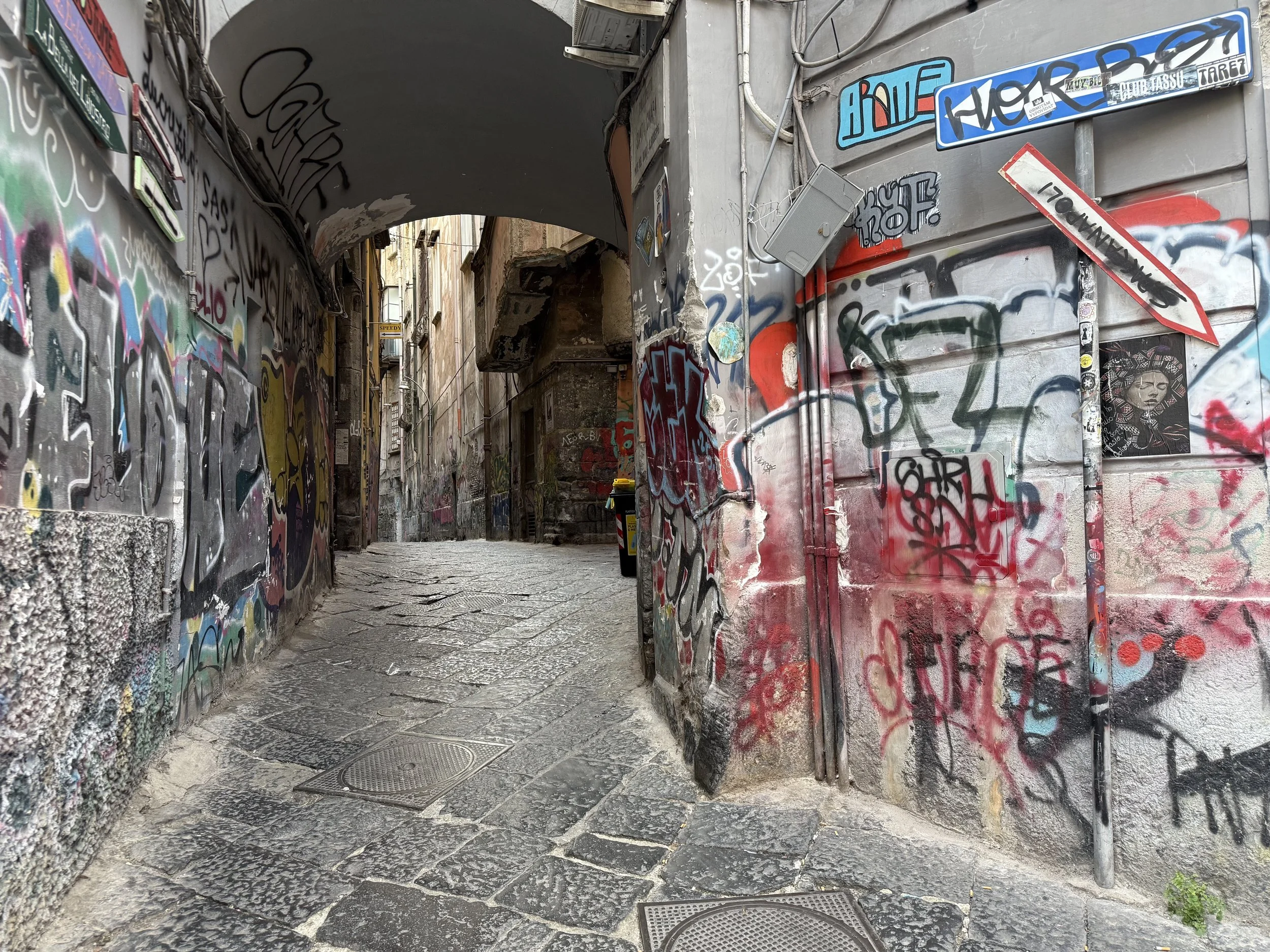 Narrow alleyway with stone paving, graffiti-covered walls, overhead arch, and a small balcony in the distance.
