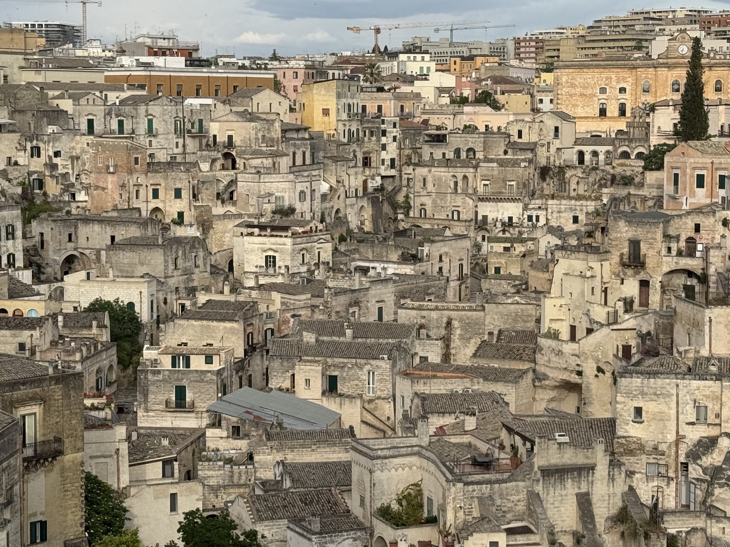 A cityscape featuring densely packed old stone buildings with terracotta and flat roofs, some with small balconies and windows, under a partly cloudy sky with construction cranes in the background.