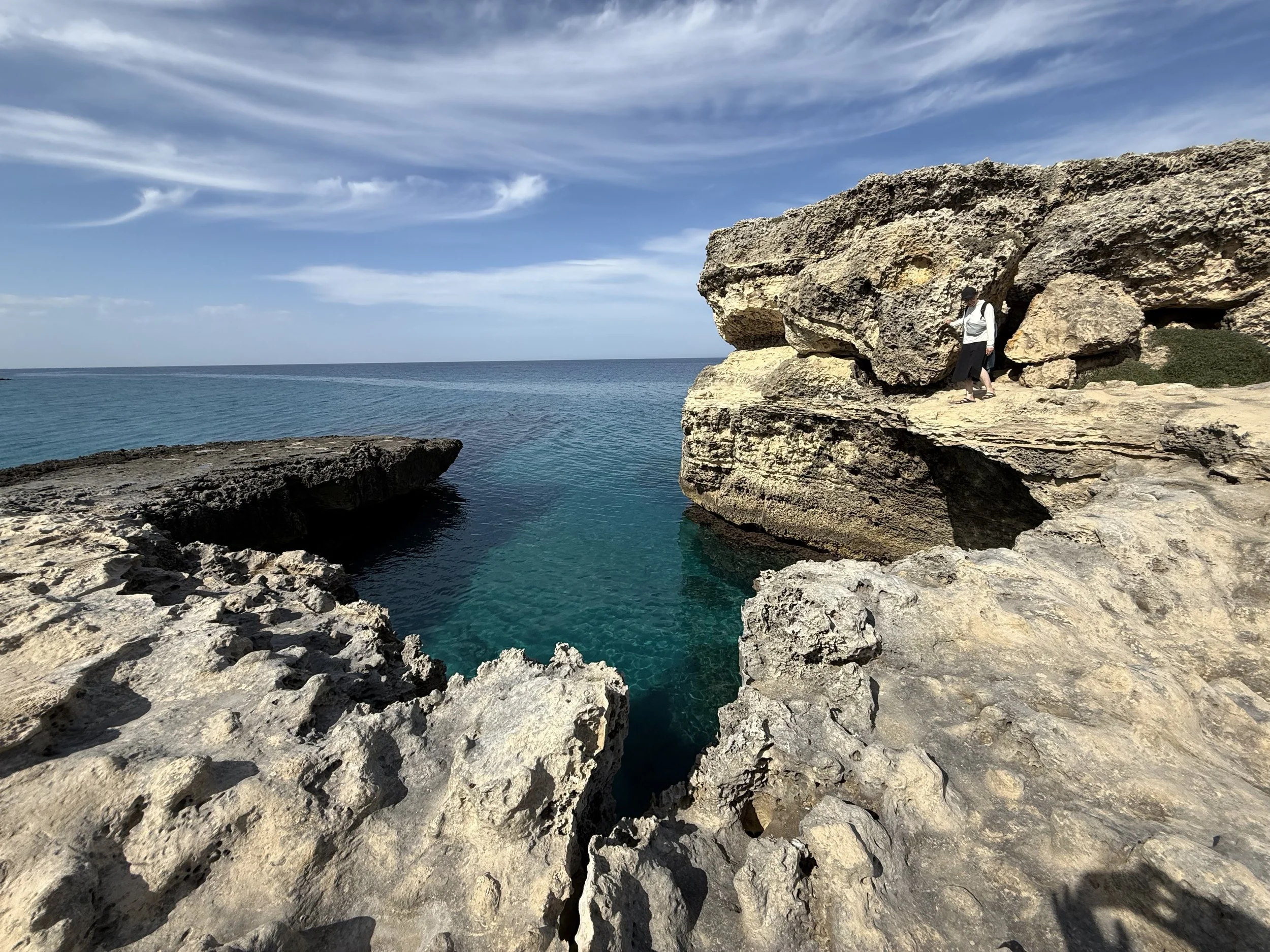 Person walking near rock formations along the ocean shoreline on a sunny day with a partly cloudy sky.