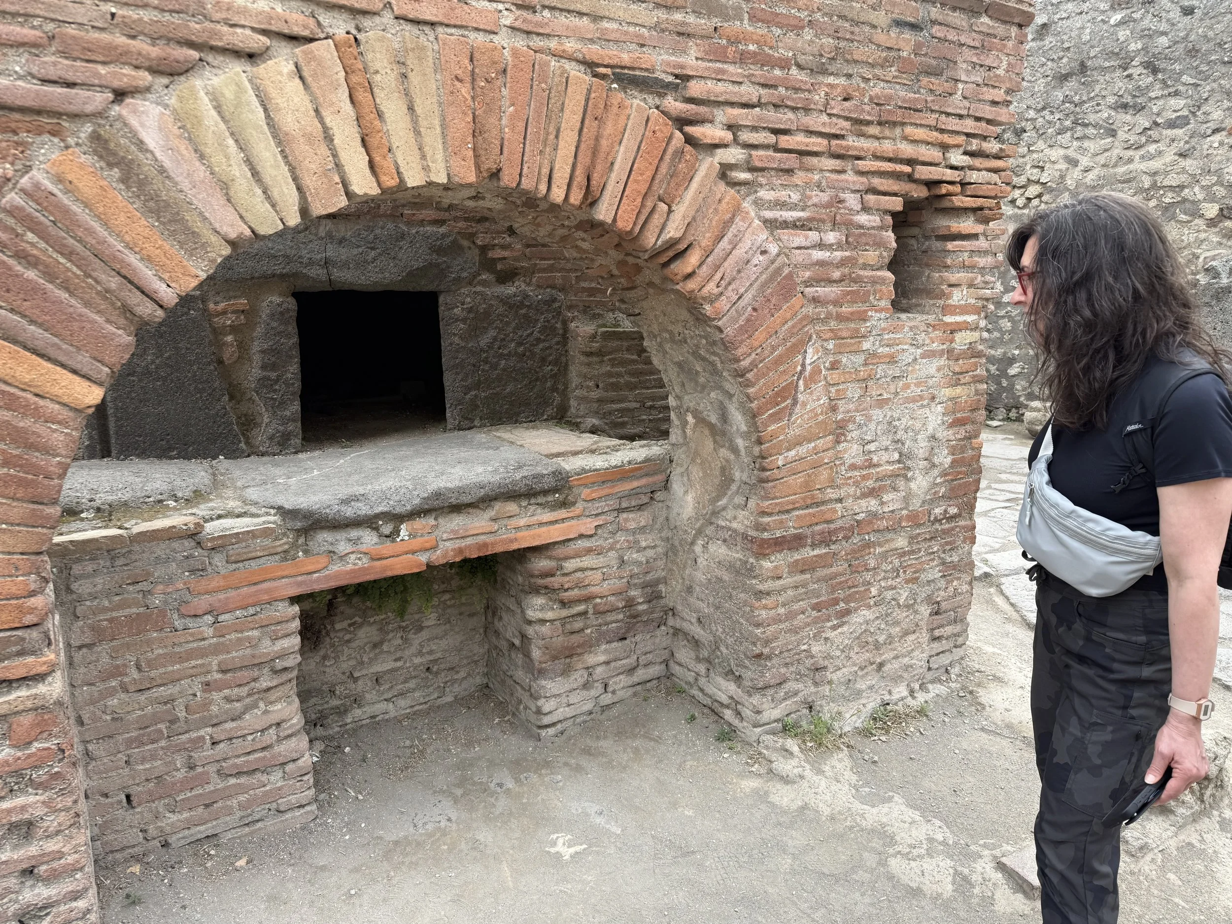 A person with long dark hair and glasses, wearing a black top and a gray bag, standing near an old brick oven or kiln outdoors.