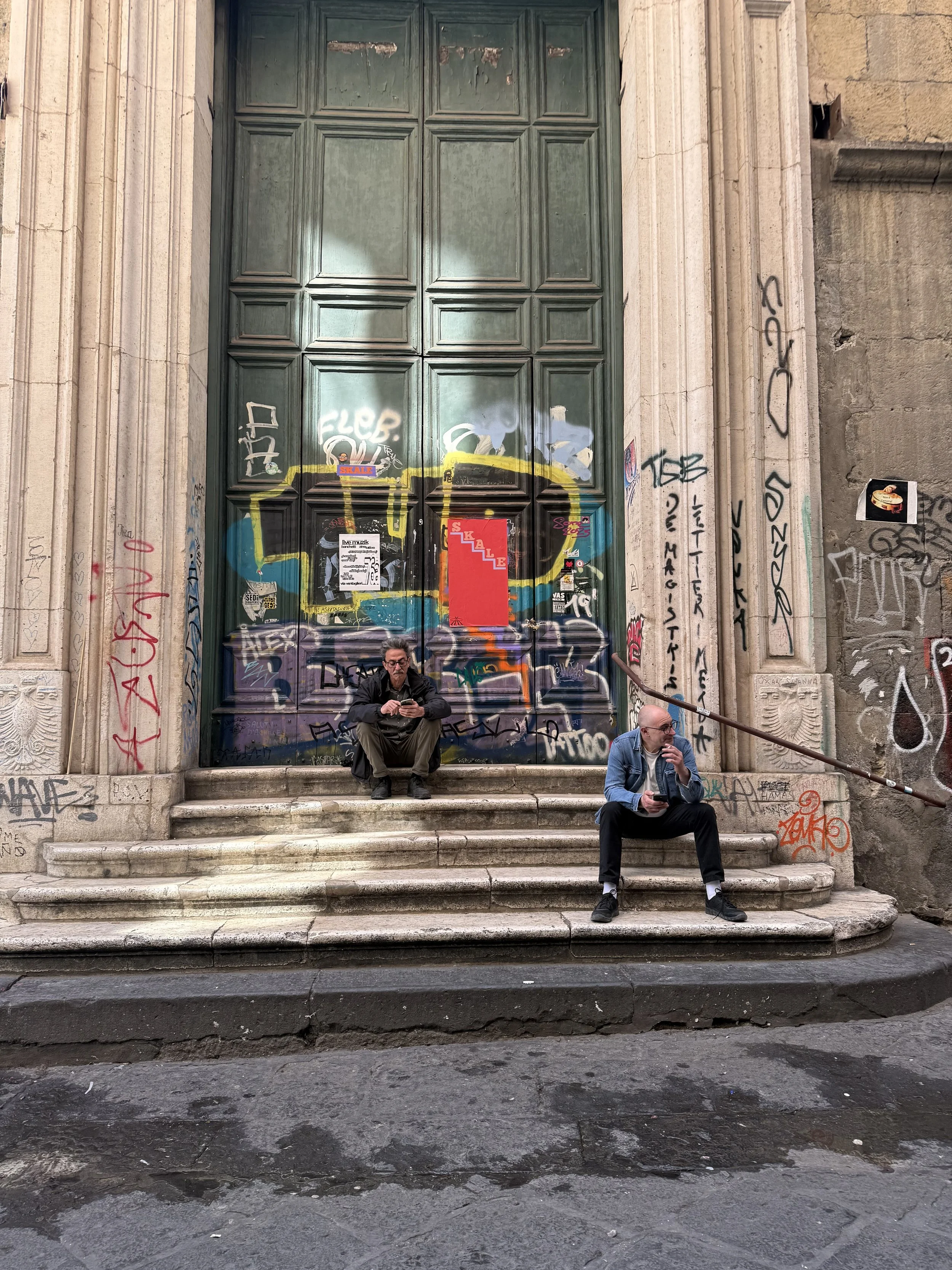 Two men sitting on the steps of a large, graffiti-covered door at a historic building. One man is using a smartphone, the other is smoking a cigarette.
