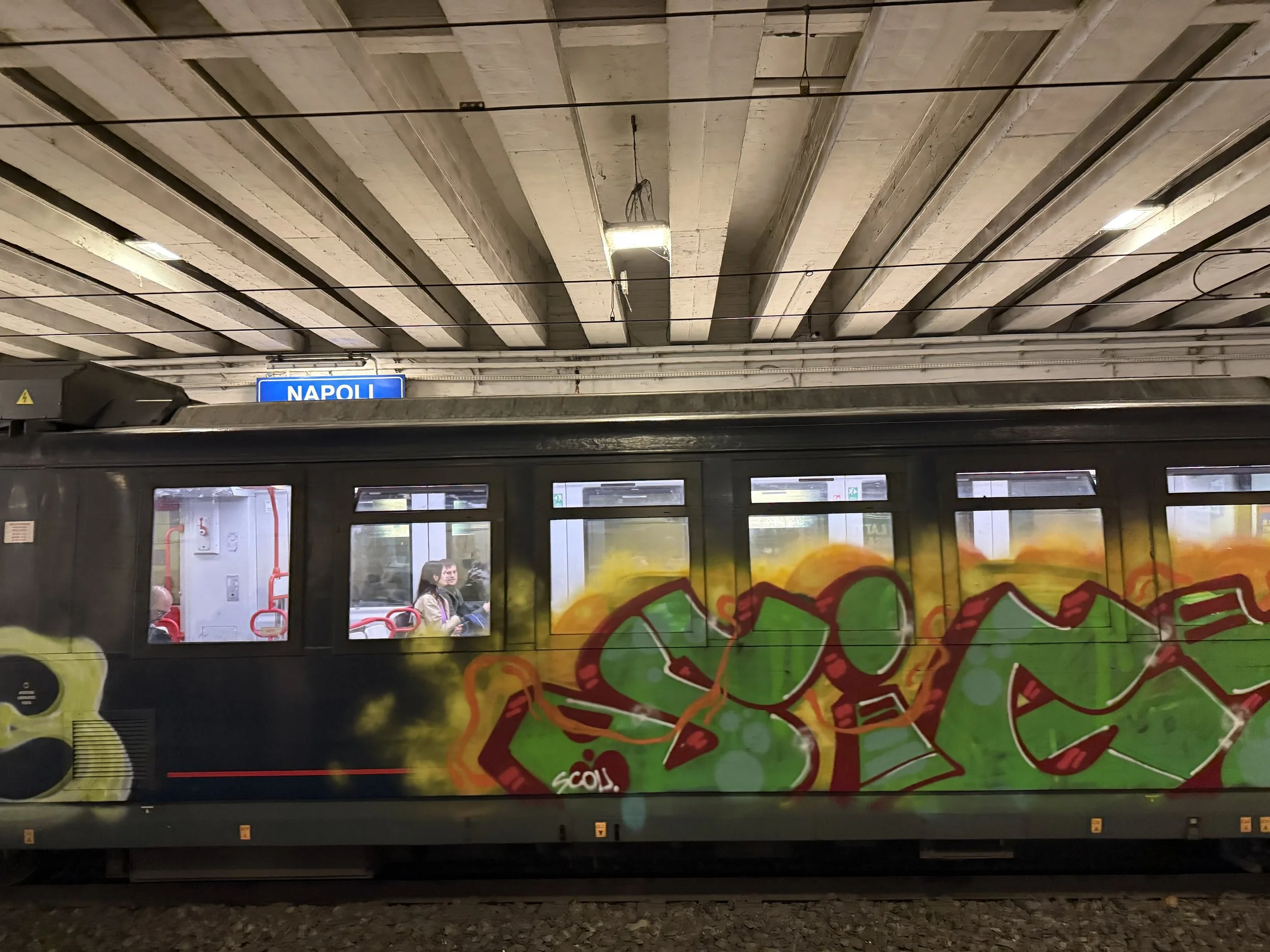 A subway train stopped at a station platform, with an illustrated city map on the side of the train and a blue sign indicating 'Napoli' in the background. Passengers are visible inside the train.