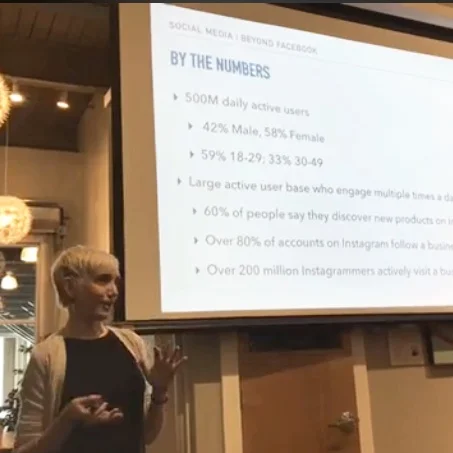 A woman stands in front of a slide show presentation, giving a talk