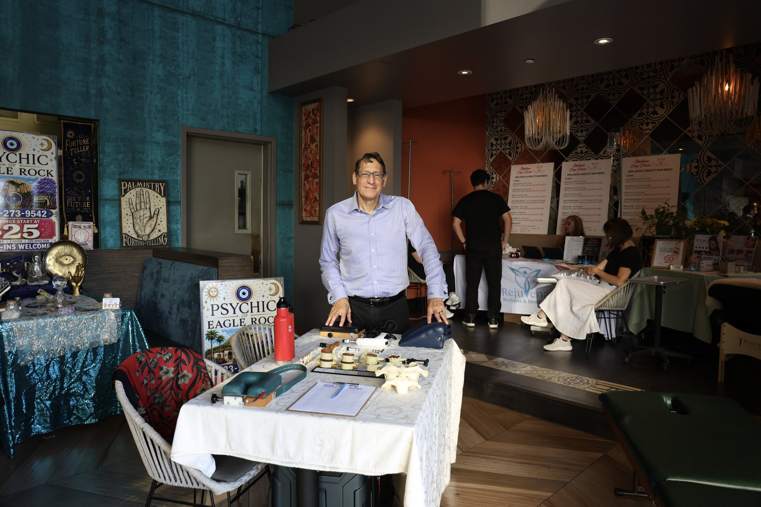 Man standing at a wellness and beauty expo booth surrounded by fortune-telling, palmistry, and chiropractic displays