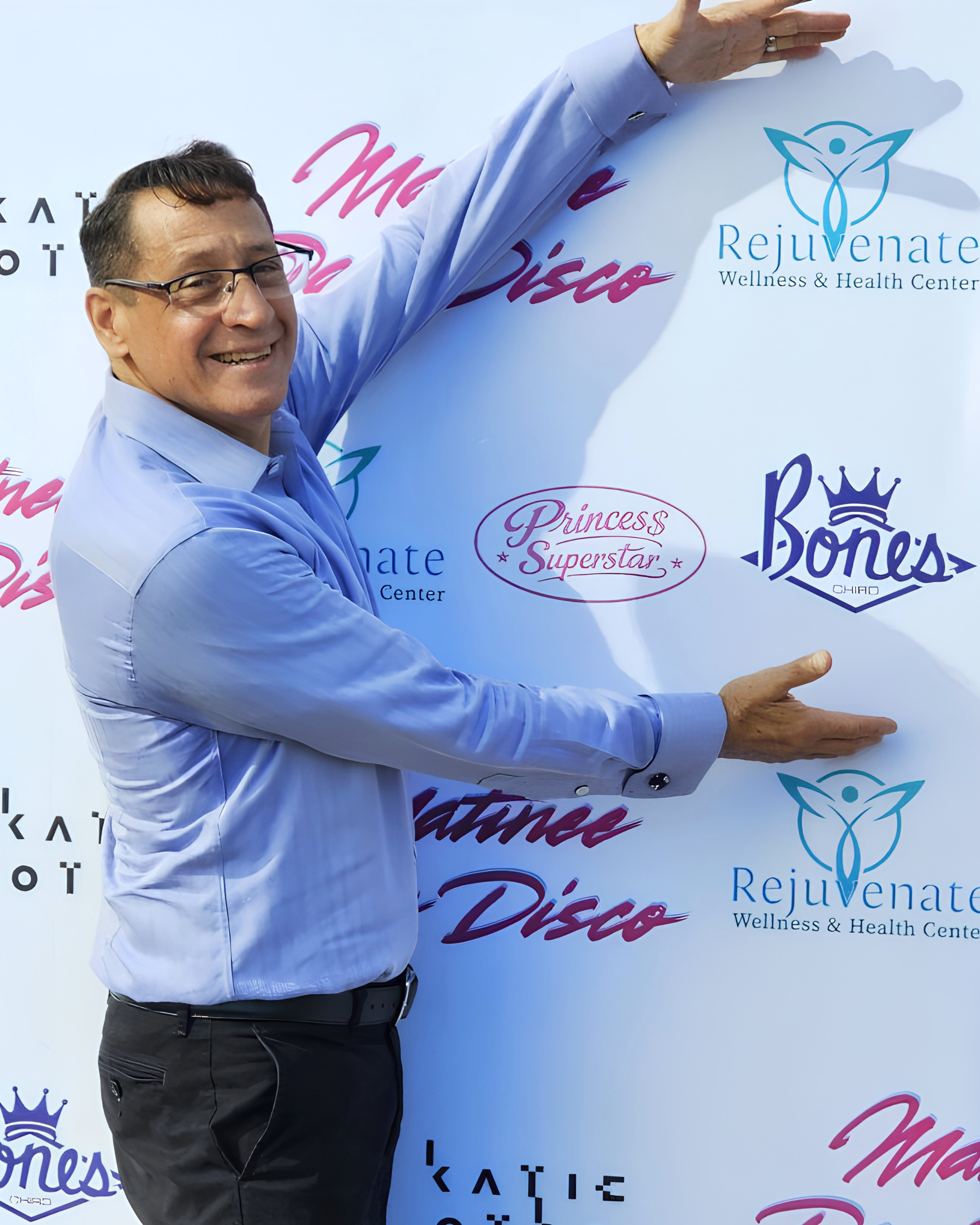 Smiling man in glasses and a blue shirt posing with outstretched arms in front of a Rejuvenate Wellness & Health Center event backdrop