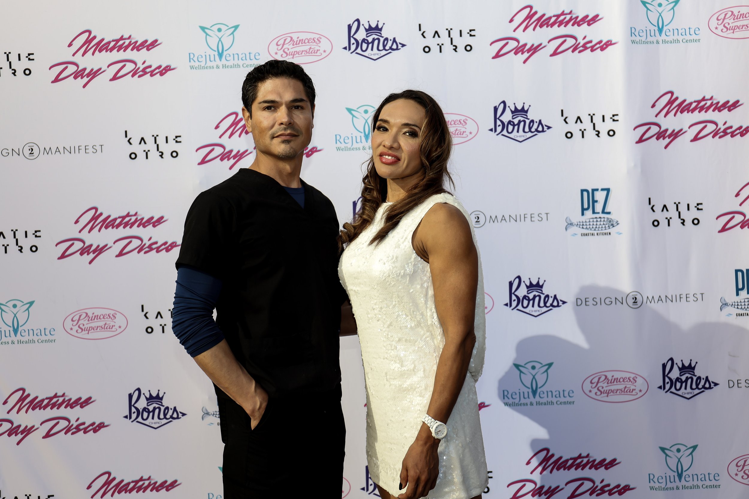 Man and woman posing at a Matinee Day Disco event in front of a sponsor-filled step-and-repeat backdrop