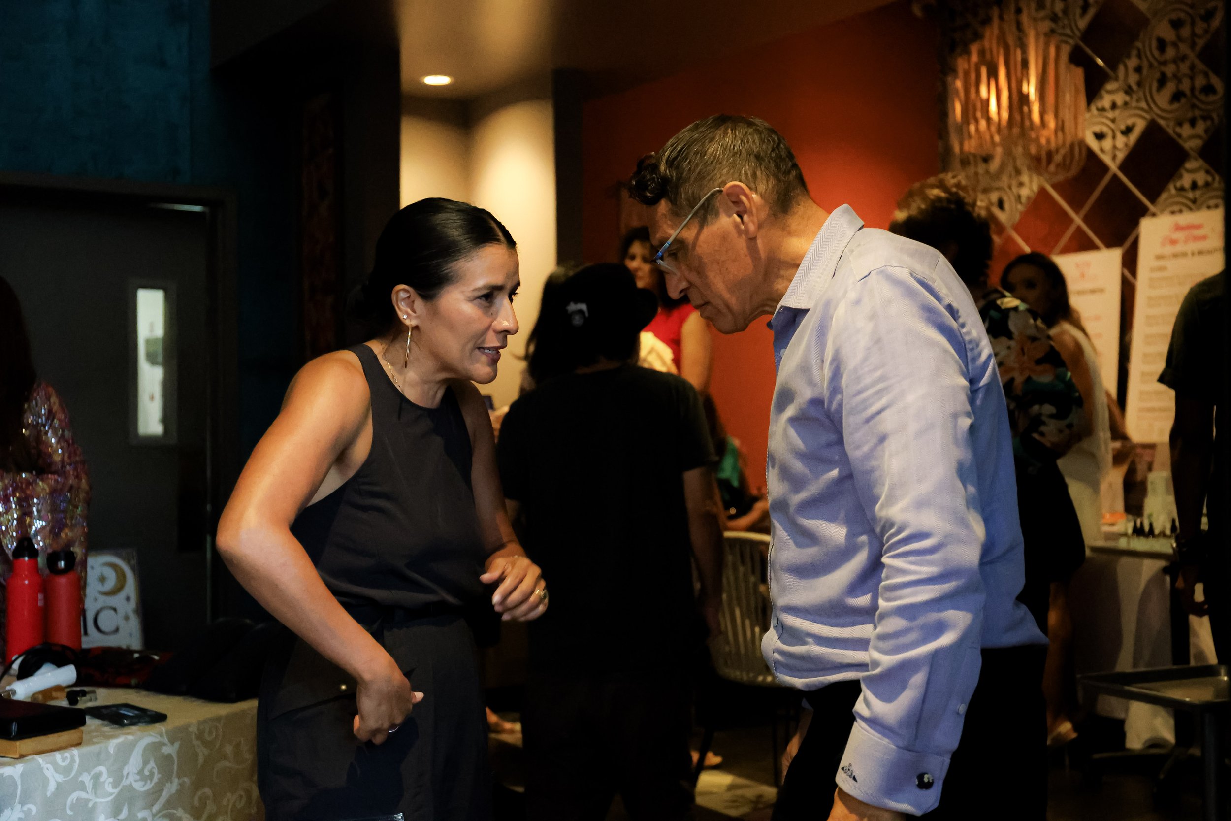 Man and woman conversing at a warmly lit indoor social event with other guests in the background