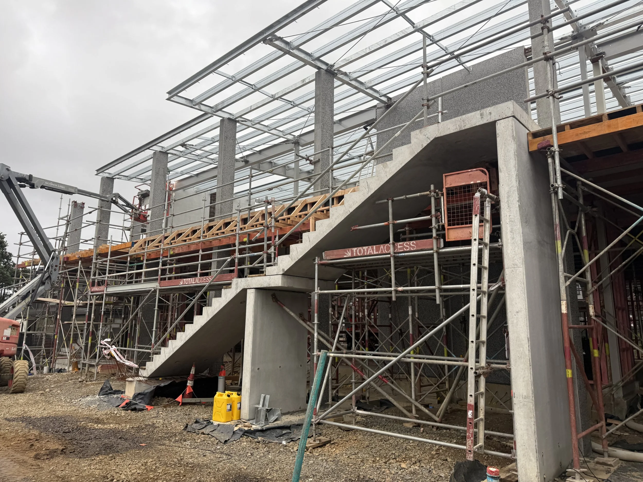 Construction site of a building with scaffolding, partially completed concrete stairs, and a glass roof structure under cloudy sky.