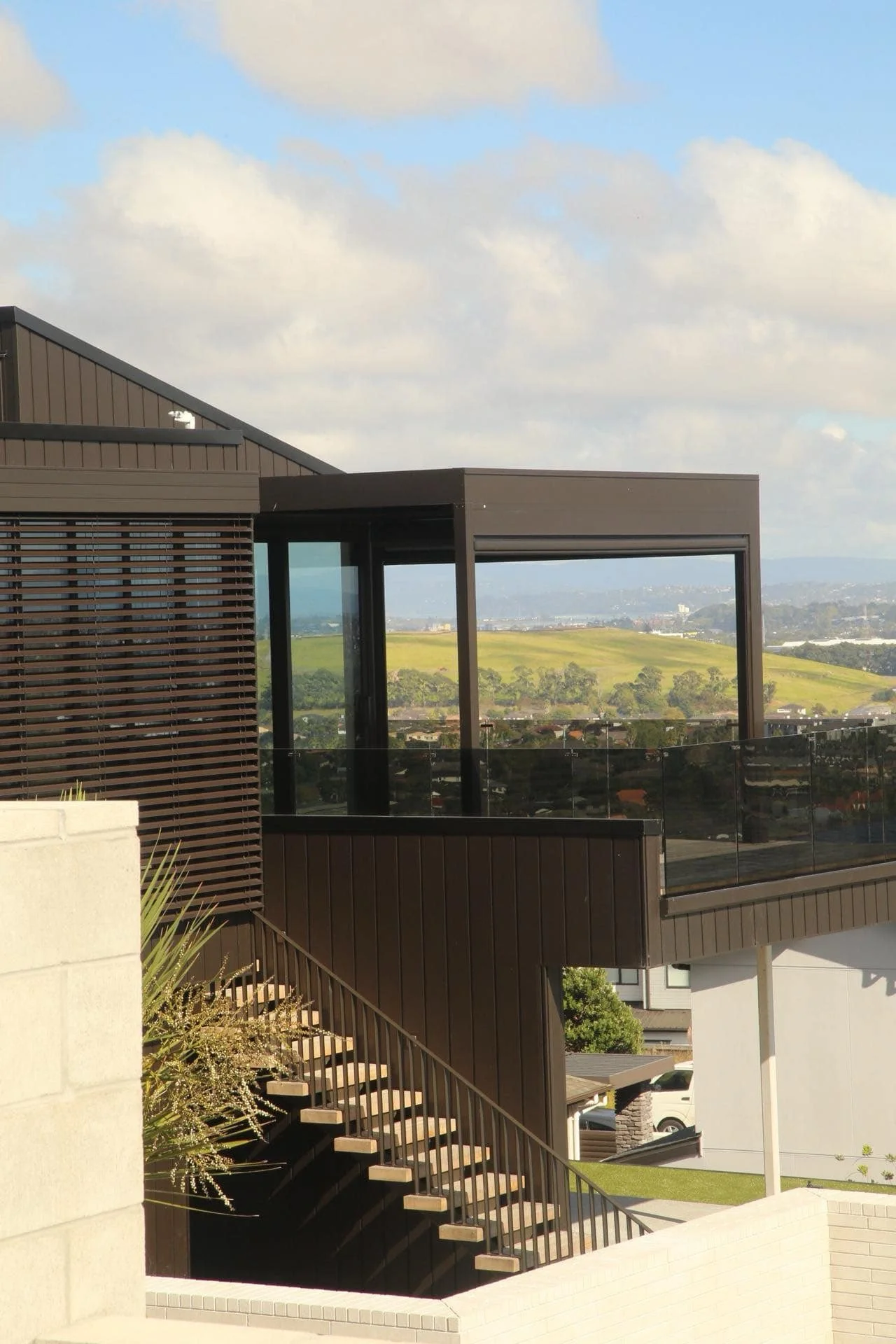 Modern building with brown siding, glass balcony, and outdoor staircase, overlooking a landscape of green hills and sky with clouds.