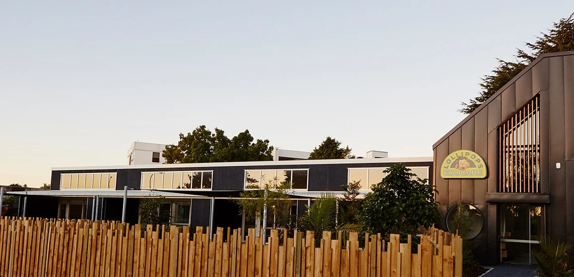 Exterior of a building with a wooden fence, large windows, trees, and a sign for Lollipops Educare.