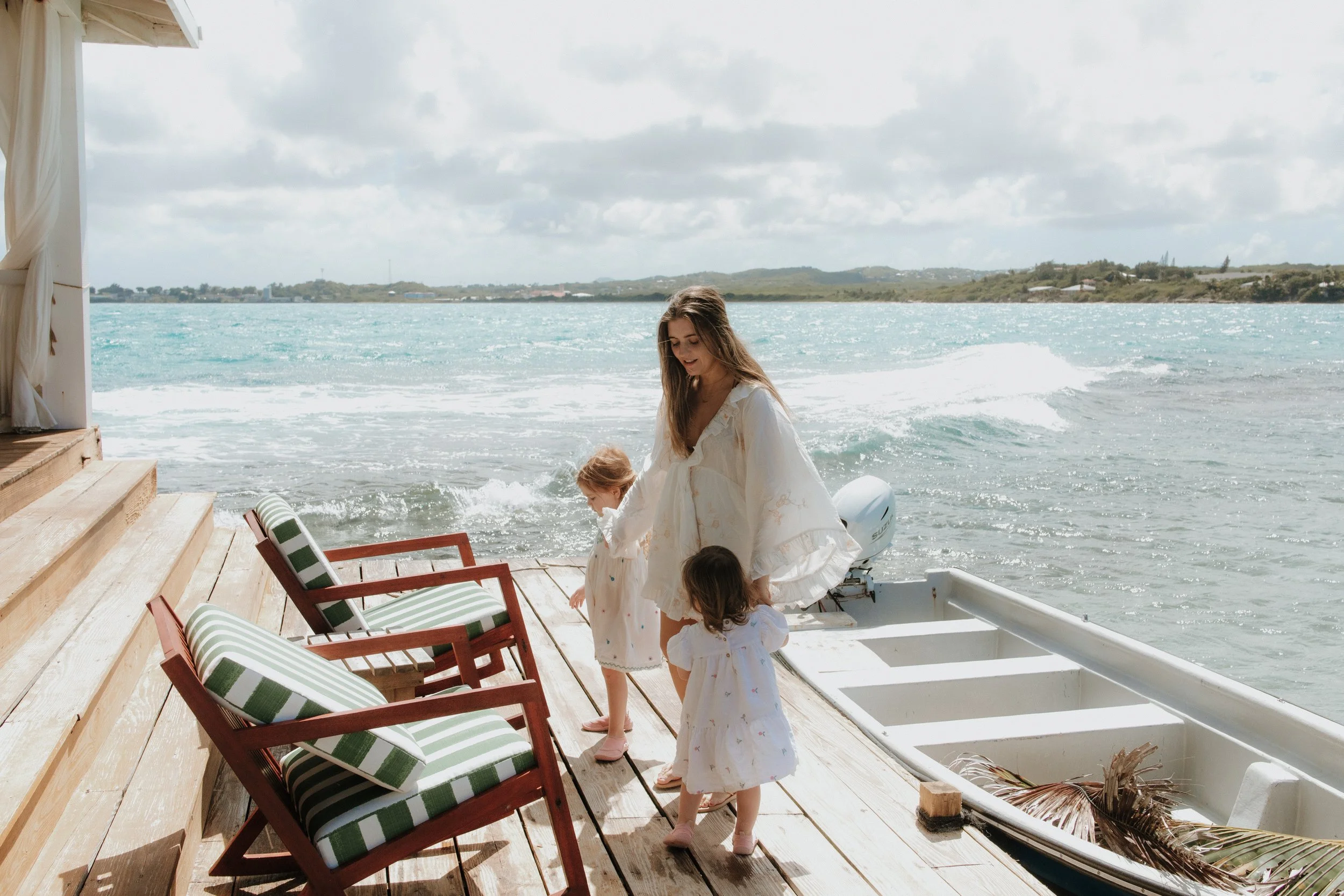 A woman with three young children on a wooden dock by the water. The woman is wearing a white dress, and the children are dressed in white dresses. There are three wooden chairs with striped cushions and a small boat with some dried palm leaves inside. The background shows a cloudy sky and a body of water with small waves.