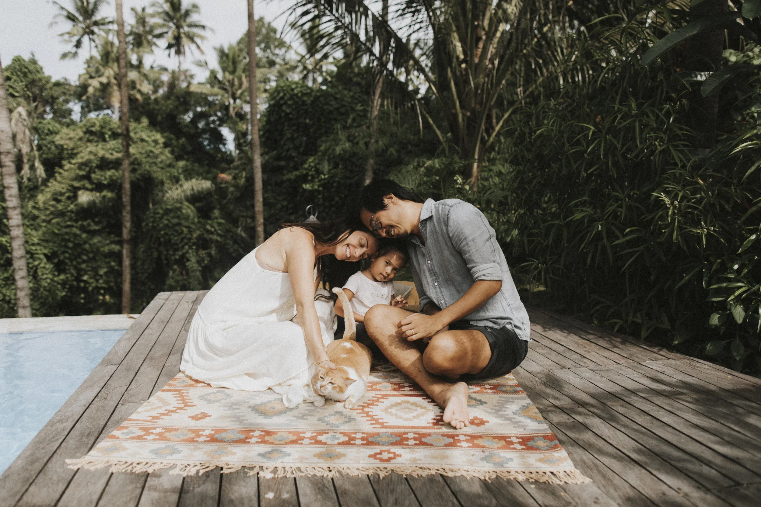 Family of three with a dog sitting on a patterned rug on a wooden deck, surrounded by lush greenery and trees.