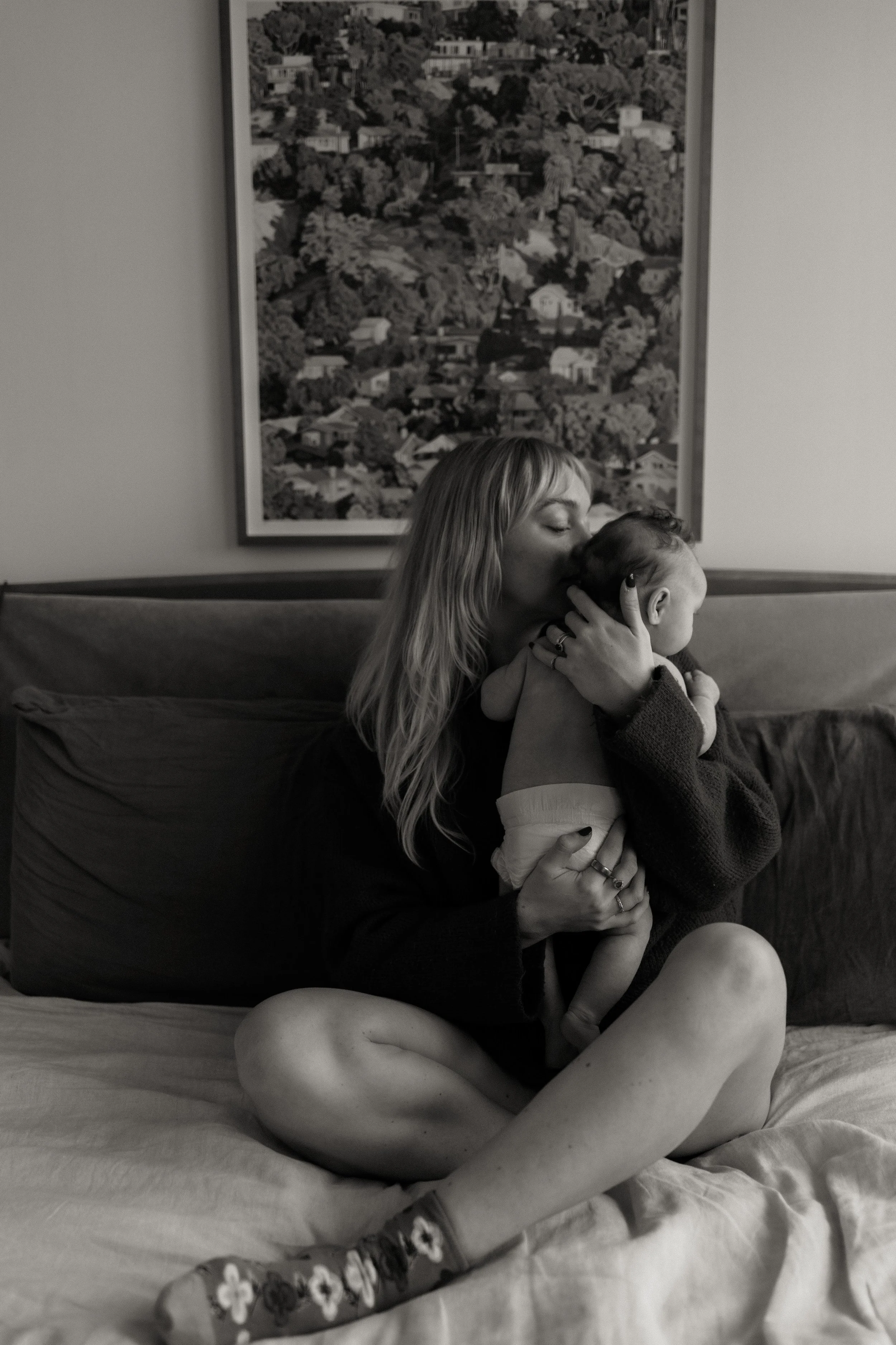 A woman with long hair sitting cross-legged on a bed, holding a crying baby and kissing their head. Behind her, there is a large framed black-and-white photograph of a hilly neighborhood.