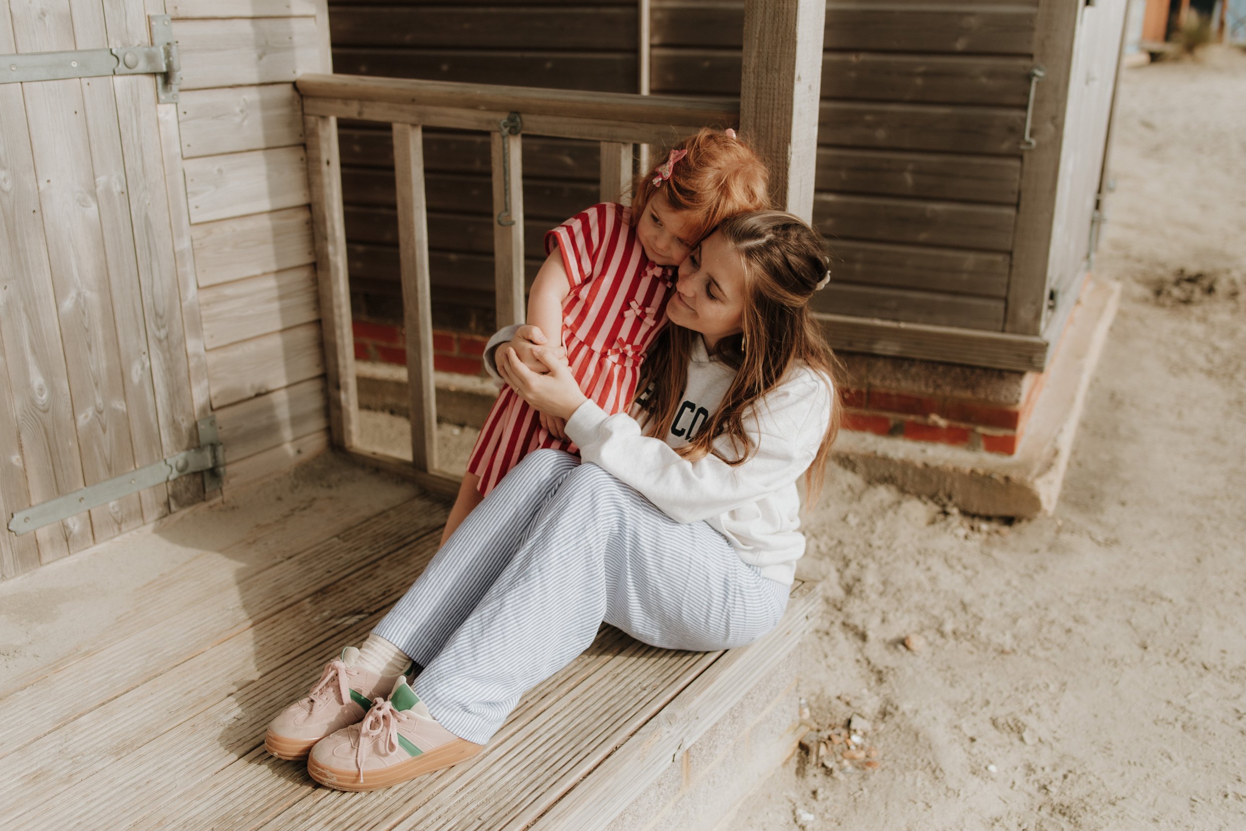 A young girl in a red and white striped dress hugging a woman seated on a wooden porch, with both their heads touching affectionately.