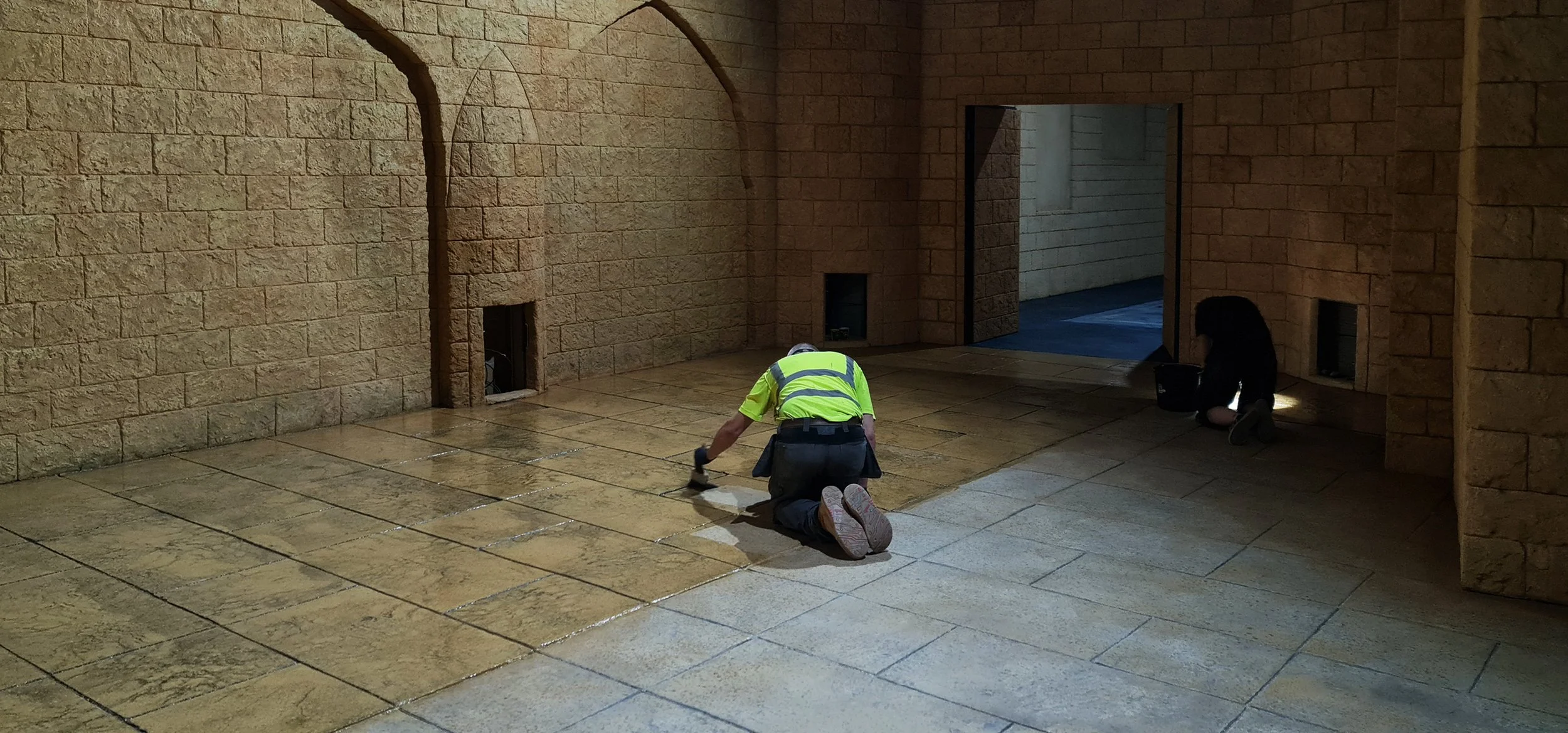 Shows two people doing the final sealant layer on a custom stamped concrete floor created and painted to emulate in keeping medieval tile to be in keeping with the carved themed and painted plaster walls of the area.