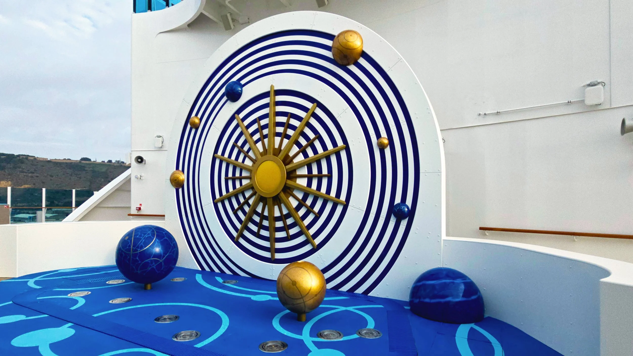 Interactive water feature built for the Star Princess cruise ship. It is a large metal sundial with fake planets painted in vibrant blues and golds with a compass like sun in the centre.