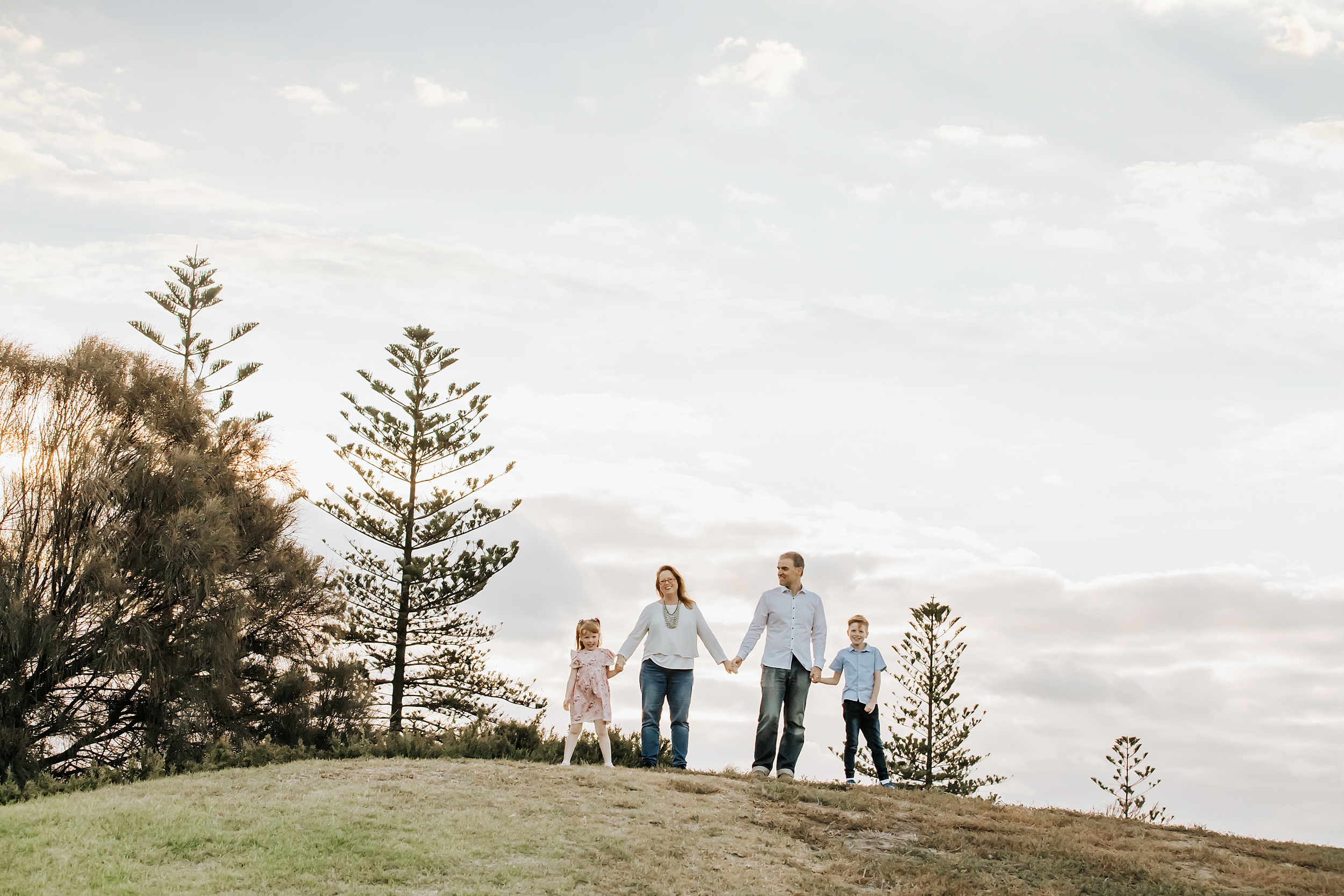 A family photo featuring a family of four standing on top of a hill all holding hands.  Taken by a professional photographer in Melbourne