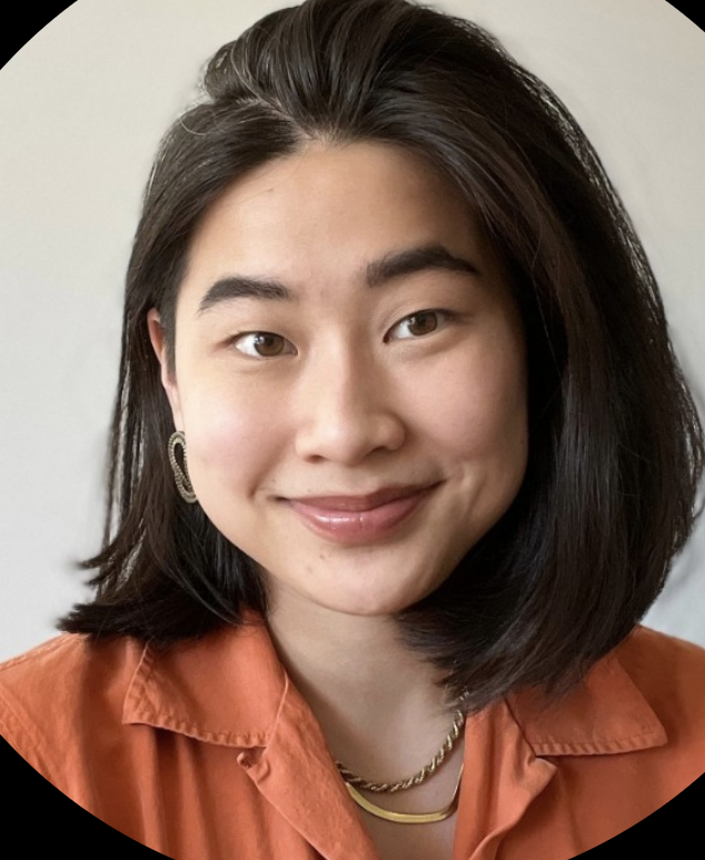Close-up portrait of a young woman with shoulder-length black hair, wearing gold jewelry and an orange shirt, smiling at the camera against a plain background.