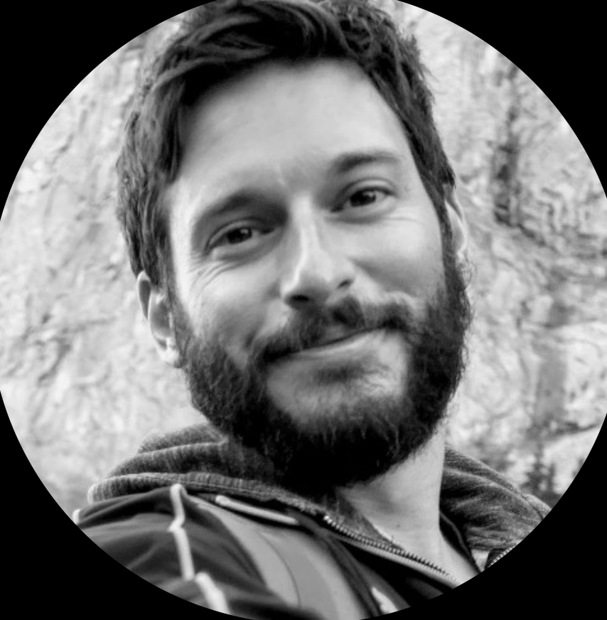 Close-up black-and-white photo of a man with a beard and short hair smiling outdoors, with a rocky background.