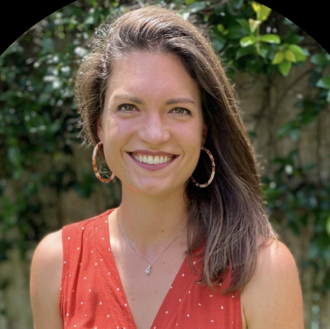 A woman with long brown hair smiling outdoors, wearing a red sleeveless top with white dots, hoop earrings, and a necklace, with green foliage in the background.