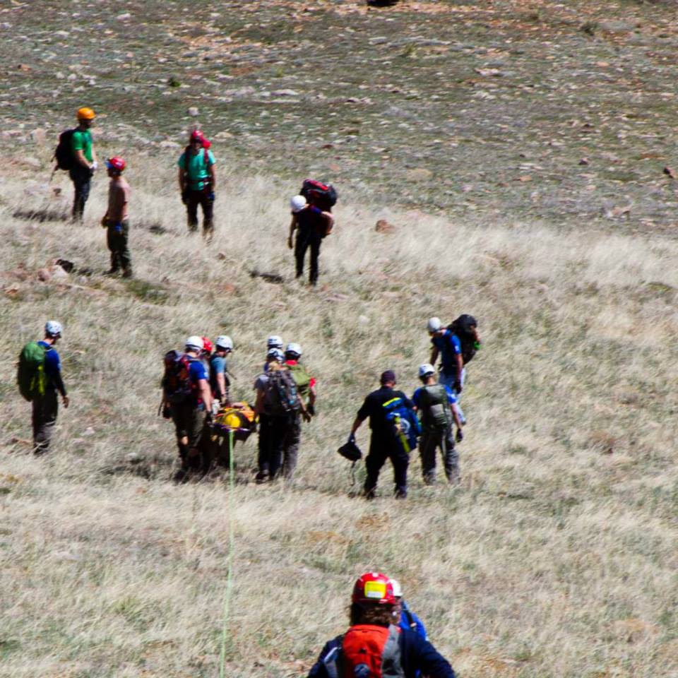 Patient with yellow helmet in litter is being lowered to the bottom of the hill (away from the camera) following his rescue after a paragliding accident. 