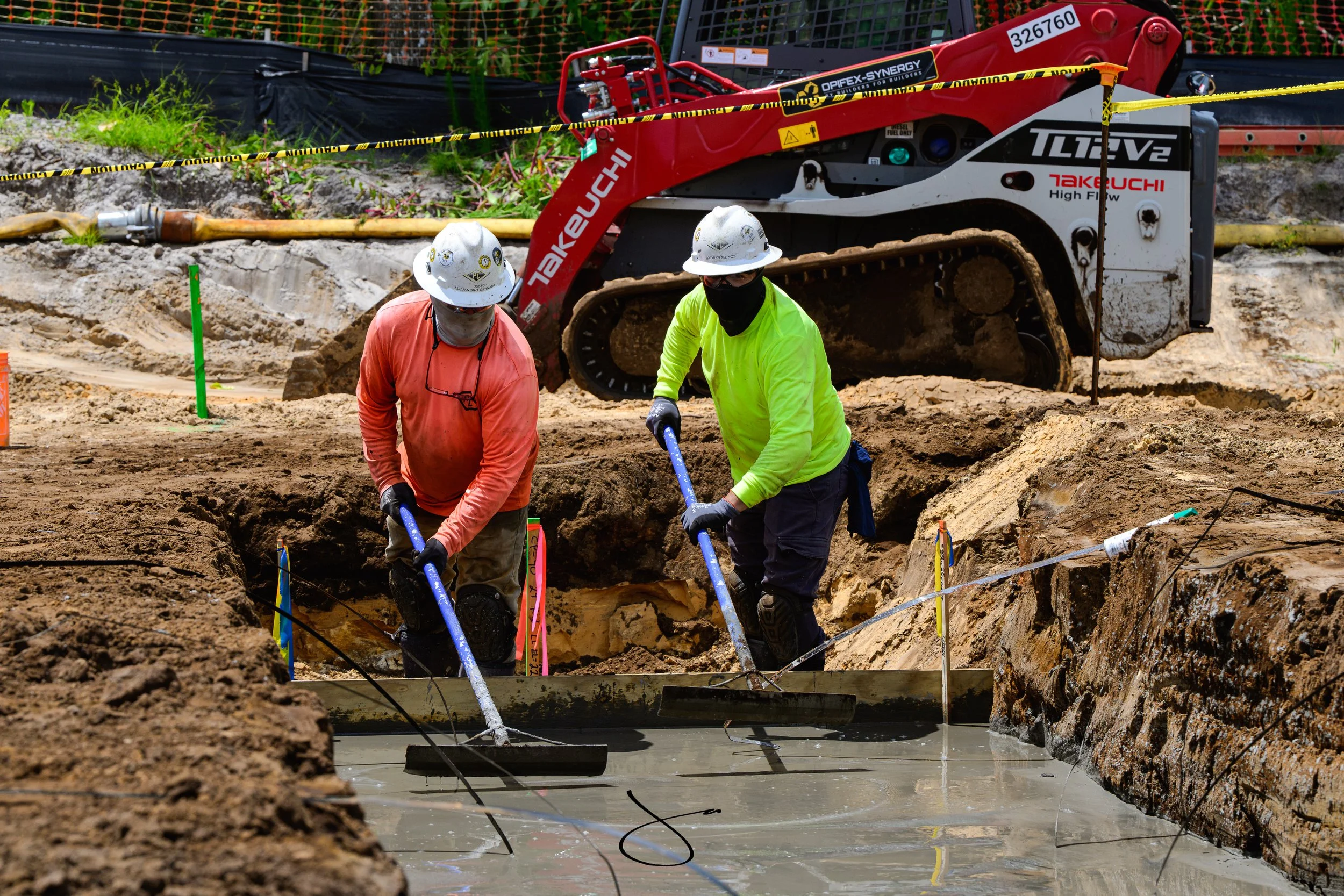 Two construction workers in neon and orange shirts wearing helmets working in a trench with a large red machinery and a yellow caution tape in the background.