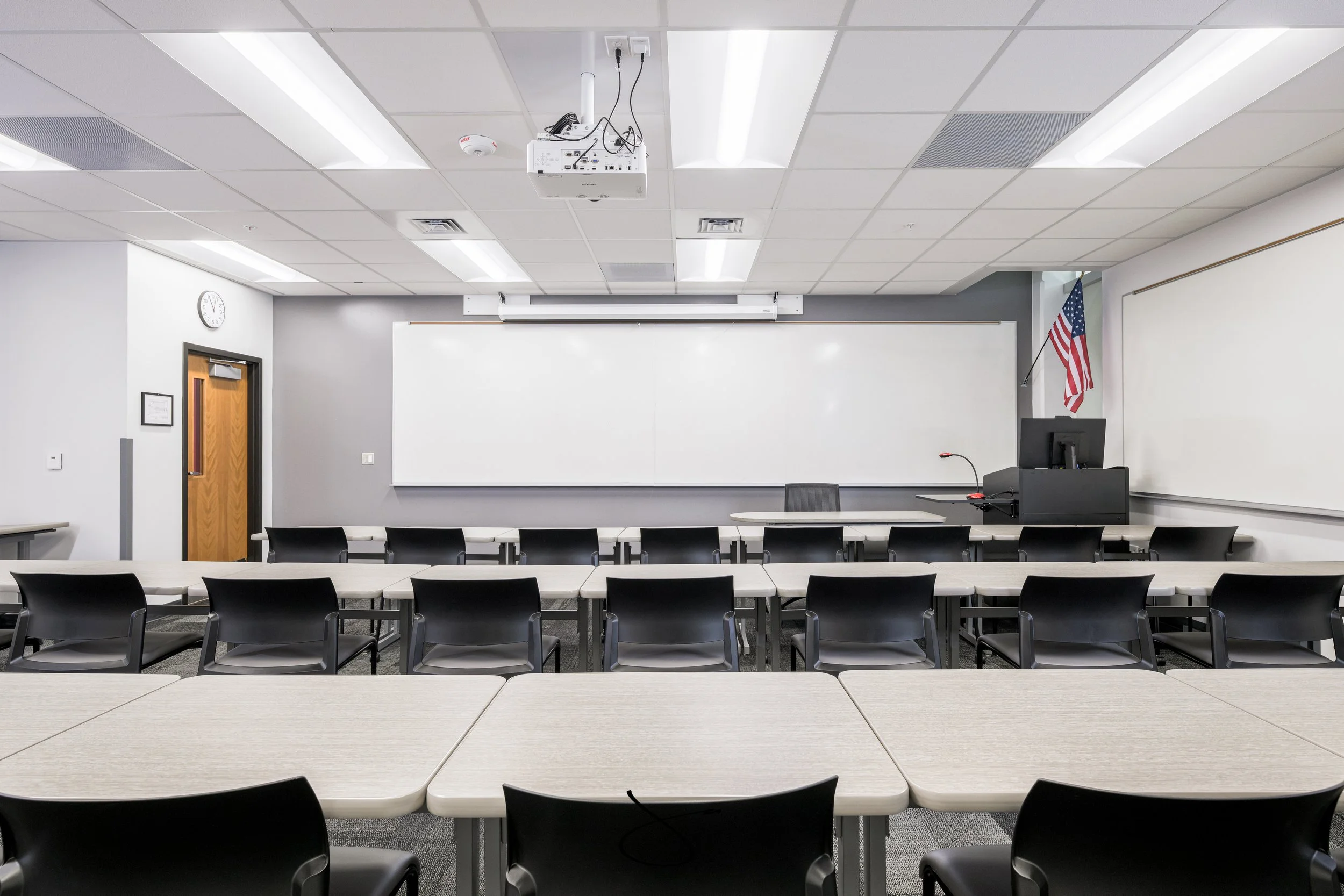 An empty classroom with long tables and black chairs, whiteboard at the front, a clock on the wall, American flag in the corner, and projector on the ceiling.