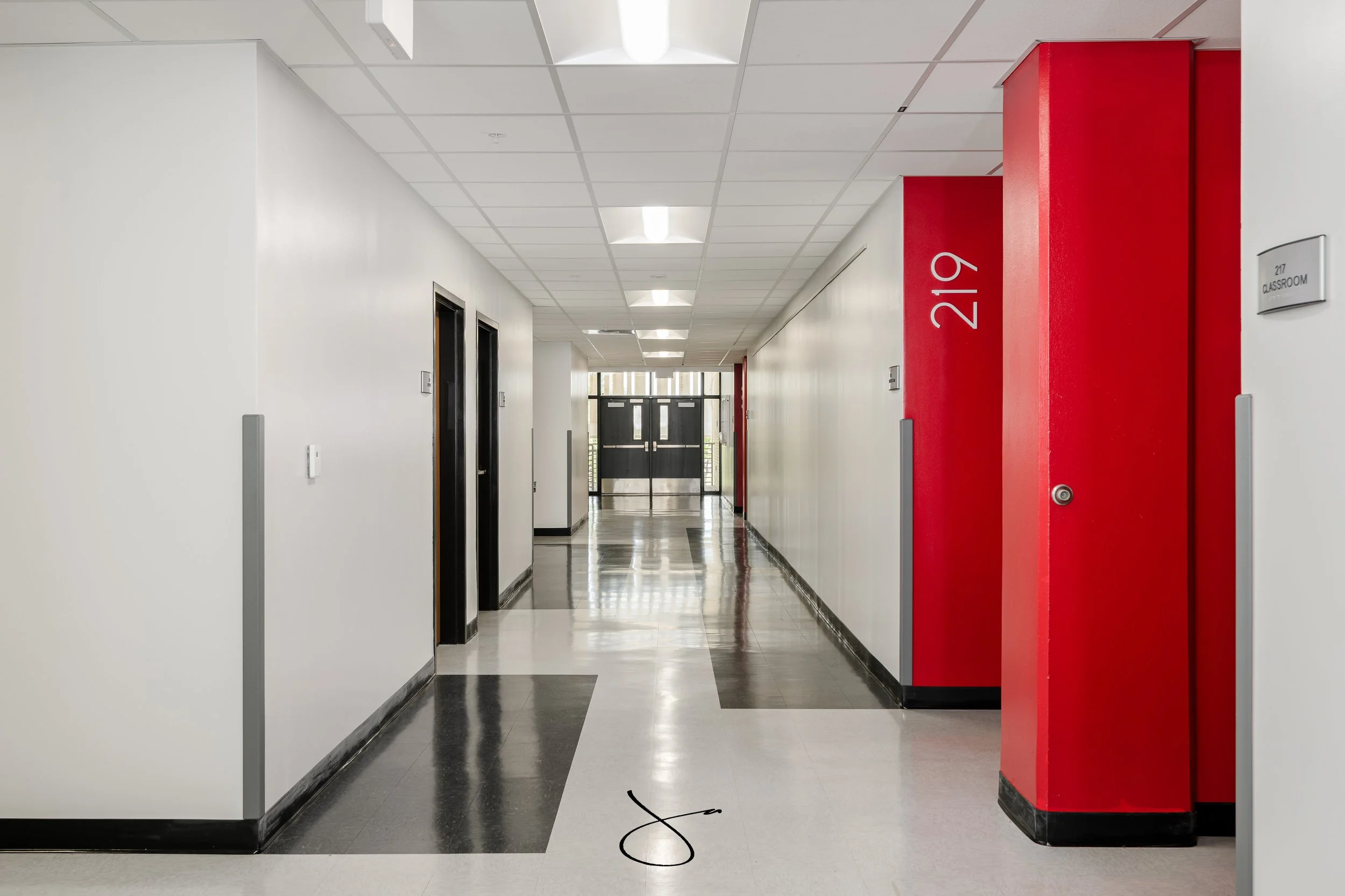 A hallway with white walls and black and white tiled flooring. Multiple doors are visible, including two elevator doors on the left and two double glass doors at the end. The ceiling has rectangular light fixtures. On the right, a red wall with the r