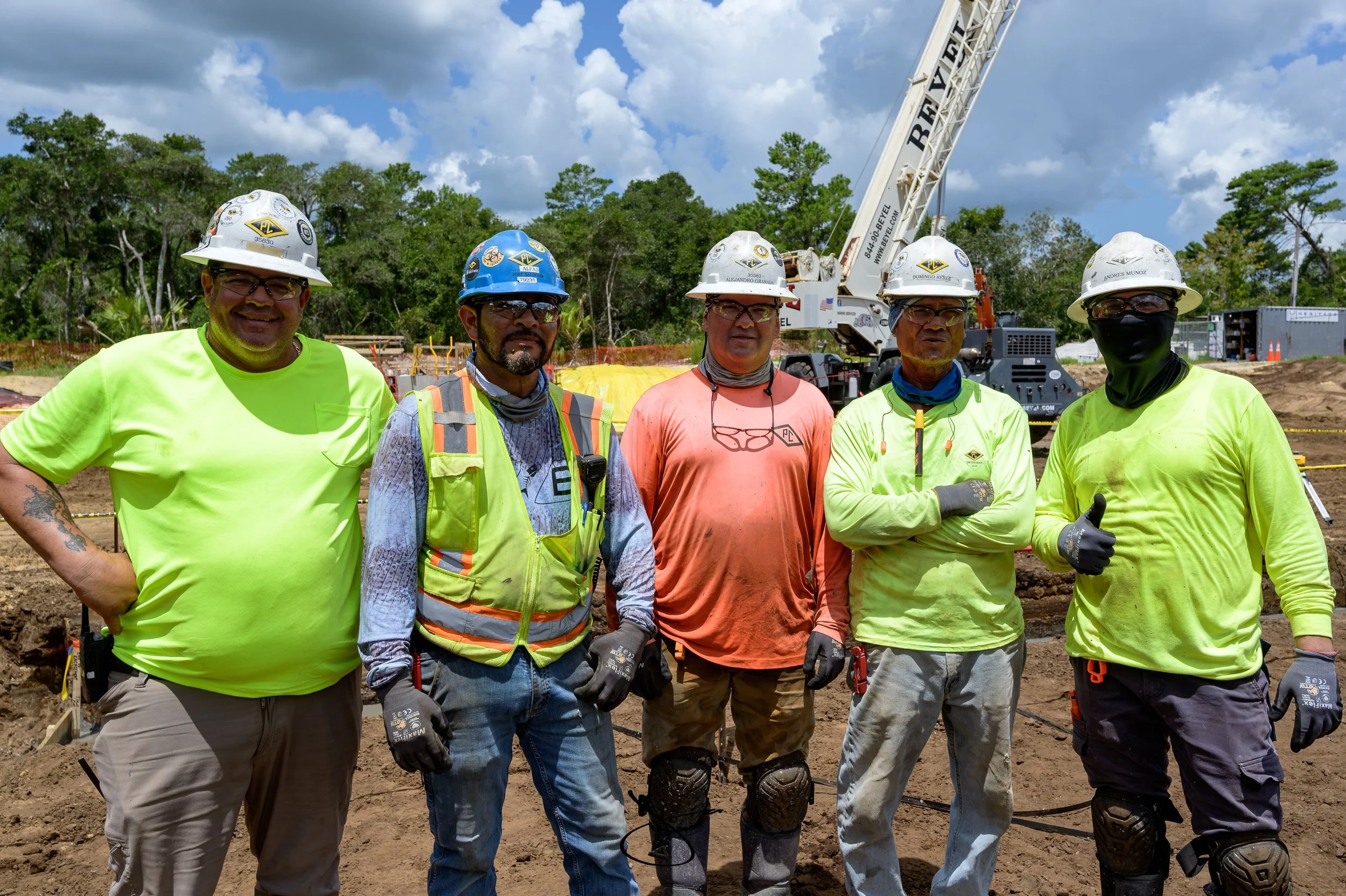 A group of five construction workers standing on a construction site, wearing safety gear including hard hats, reflective vests, and gloves. A crane is visible in the background, along with trees and a partly cloudy sky.
