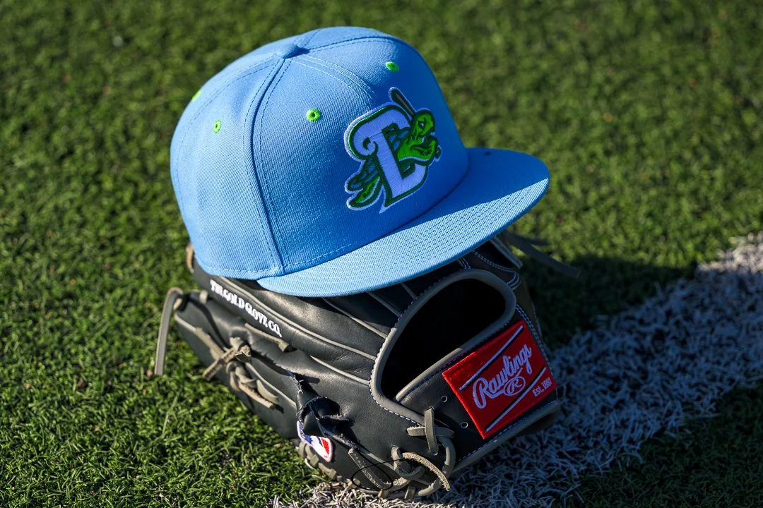 A blue baseball cap with a green insect logo rests on a black baseball glove on grass near a white line.