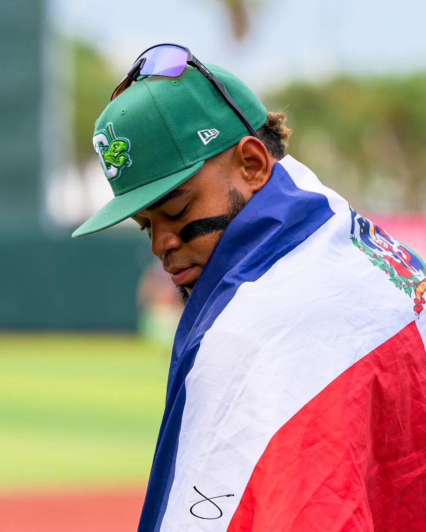 A man wearing a green baseball cap with sunglasses perched on top, a black eye black stripe, and a flag draped over his shoulders.