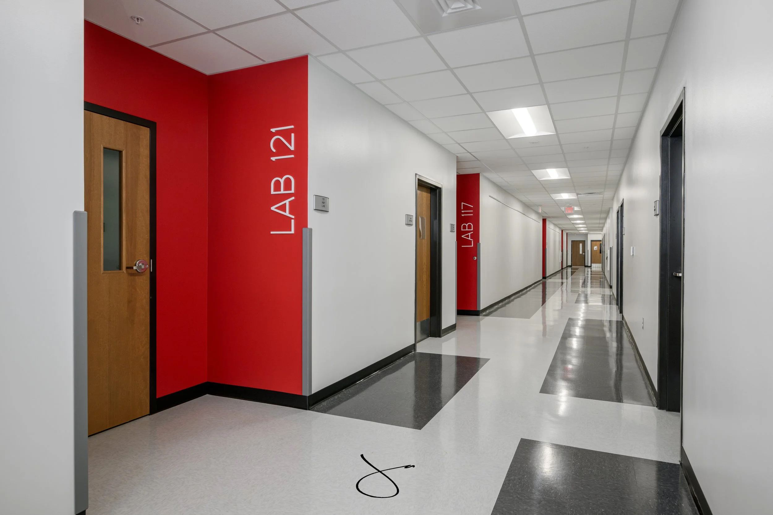 Long college corridor with white walls, a red accent wall with white door and signage reading 'LAB AB 121,' ceiling with fluorescent lights, and polished tiled floor.