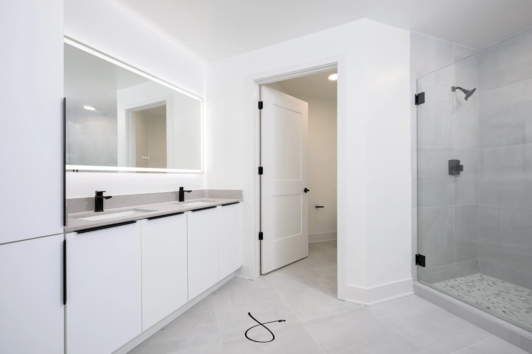 Modern white bathroom with a double vanity, black fixtures, large mirror with LED lighting, and a walk-in shower with a glass door and gray tiles.