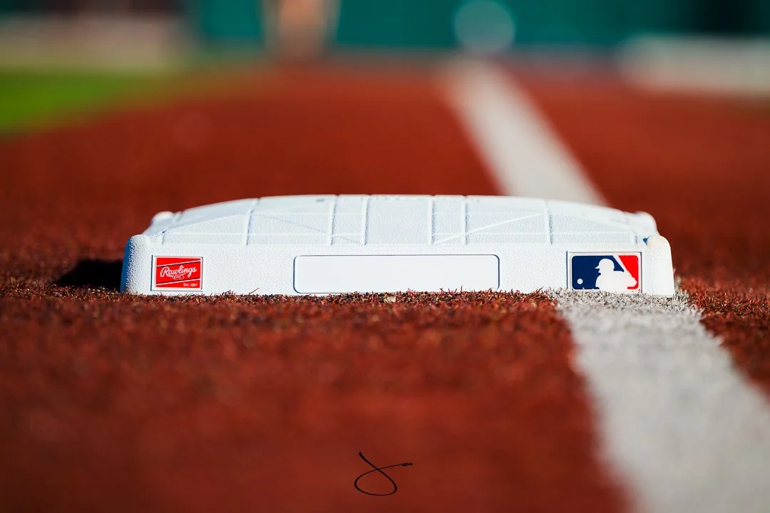 Close-up of home plate on a baseball field with a white marker featuring the MLB logo on the right and the Rawlings logo on the left. The field's red clay and white chalk lines are visible.