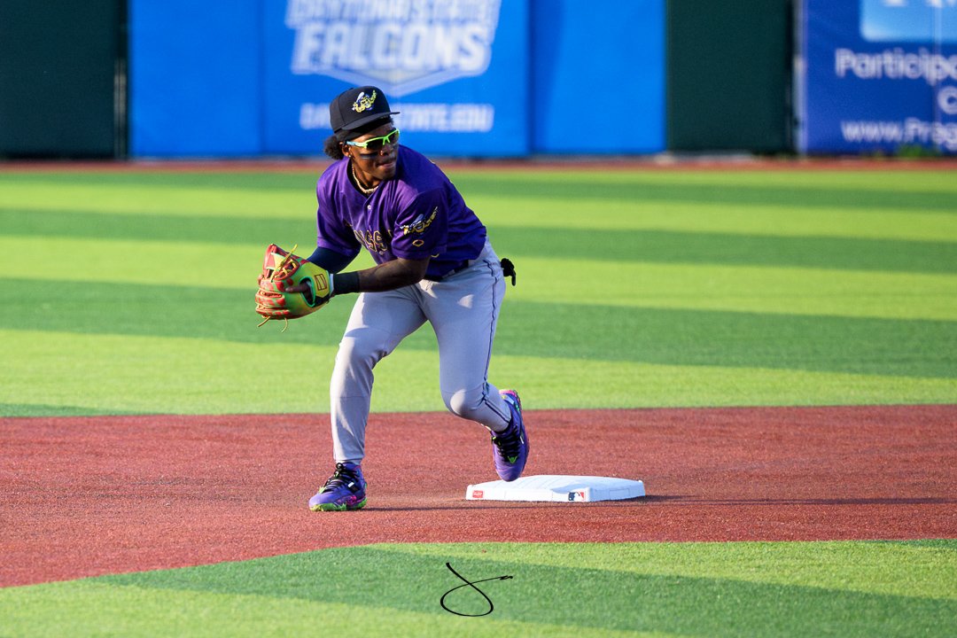 A baseball player is in a ready position on the field, wearing a purple jersey, light gray pants, and a black cap with a logo. The player is holding a baseball glove and is positioned near a base on the field.