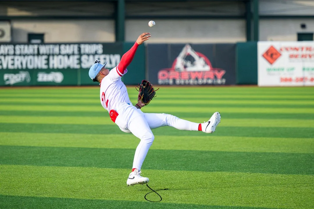 A baseball player in a white and red uniform catches a baseball with a glove in a field with green grass and advertisements in the background.