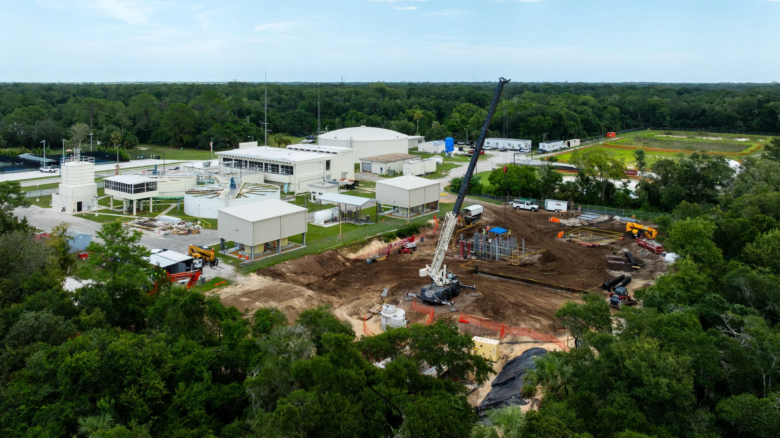 Aerial view of a construction site with work being done on the ground, surrounded by trees and infrastructure building in the background.