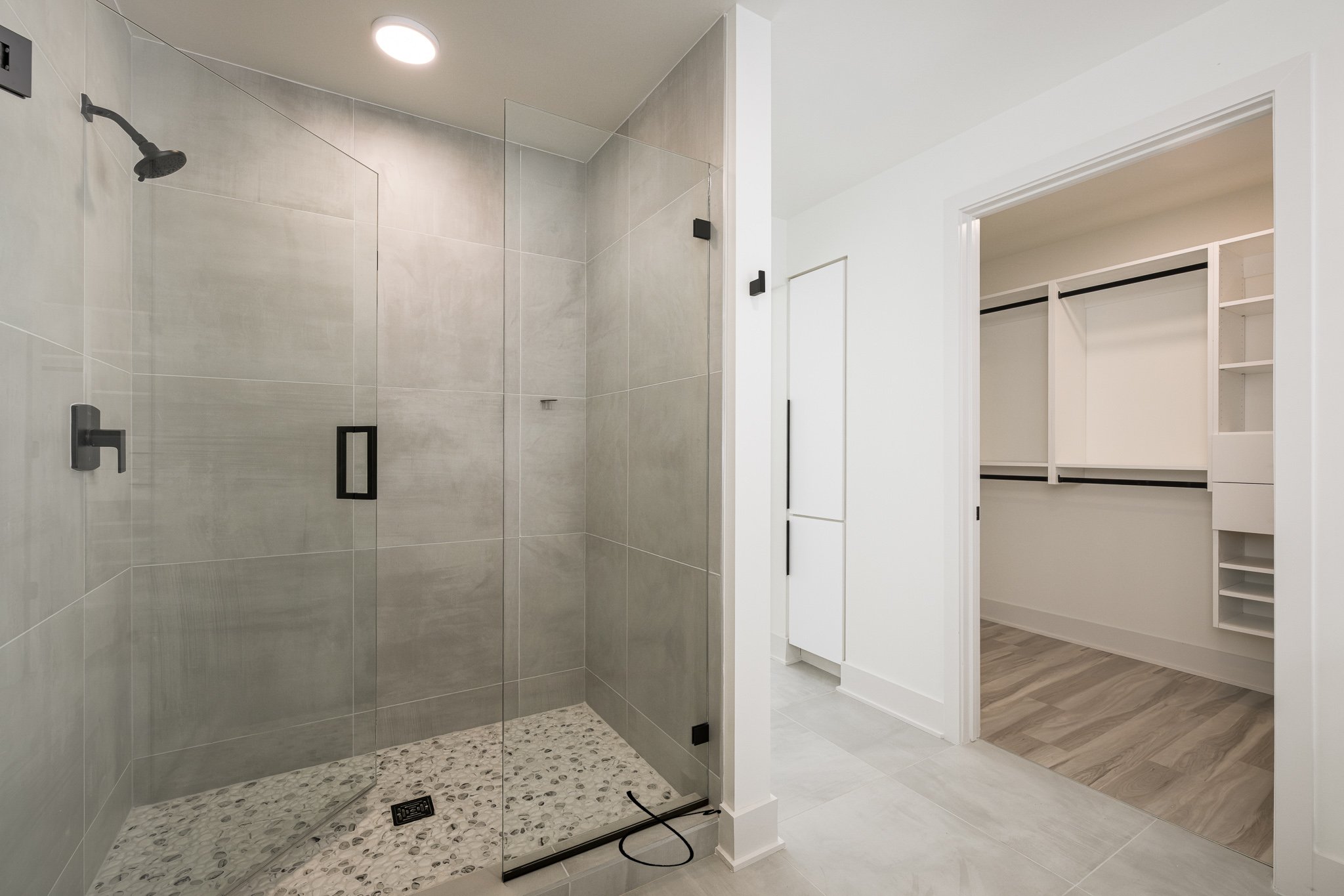 Modern walk-in shower with gray tiled walls, pebble floor, black fixtures, and a glass door. Adjacent walk-in closet with white shelving and hardwood floors.