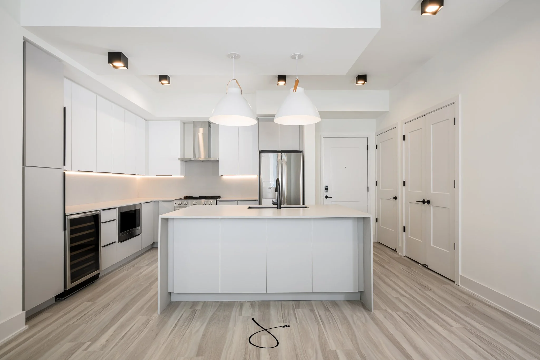 Modern white kitchen with island, stainless steel appliances, black and white lighting fixtures, and wood flooring.