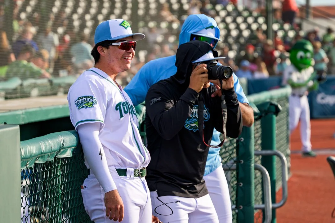A baseball player in a white uniform with teal accents and a blue cap standing near a green dugout fence, smiling and wearing sunglasses. Another person in a black jacket and black cap is taking a photo, with a third person in blue and a mascot in th