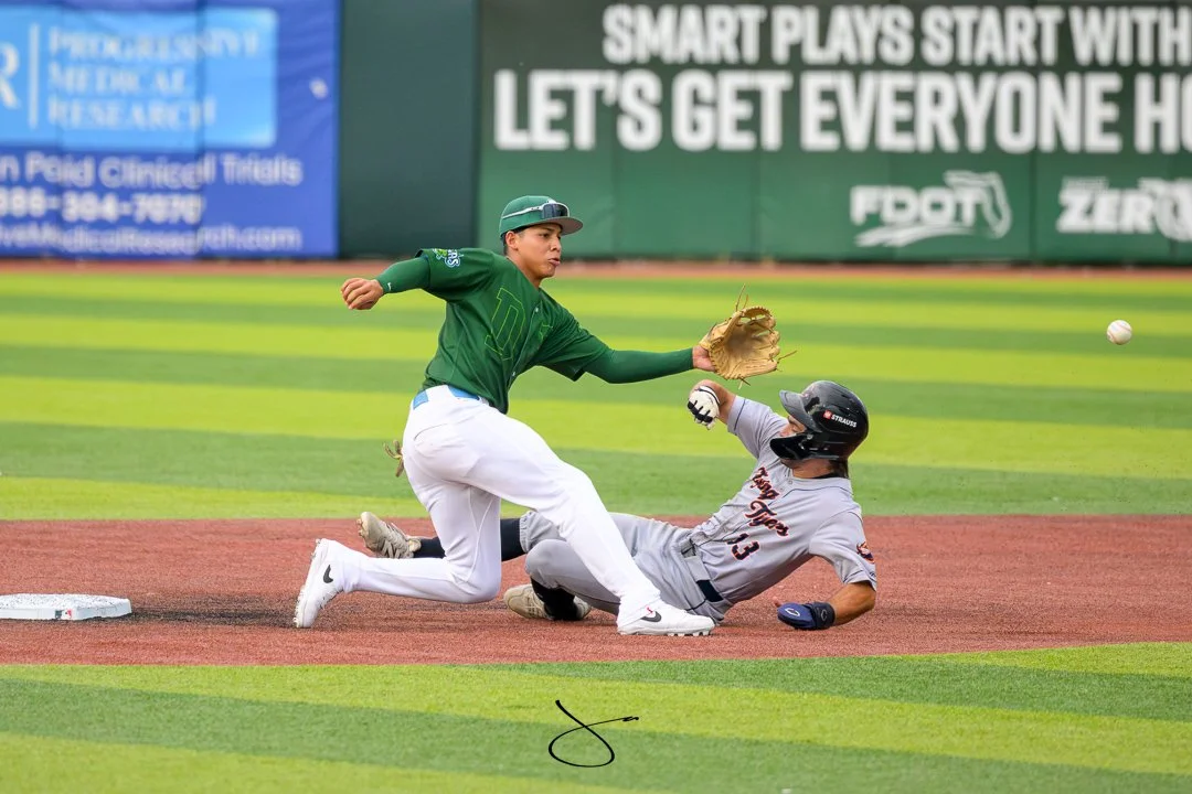 A baseball player in a gray uniform sliding into a base while another player in a green uniform attempts to tag him out, with a baseball in midair in the background.