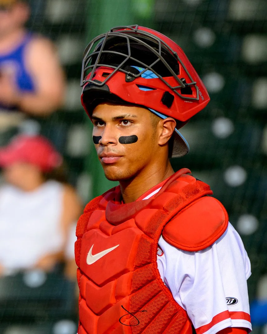 A male baseball catcher wearing a red helmet, red chest protector, and white jersey with red accents, standing on a baseball field with a focused expression.