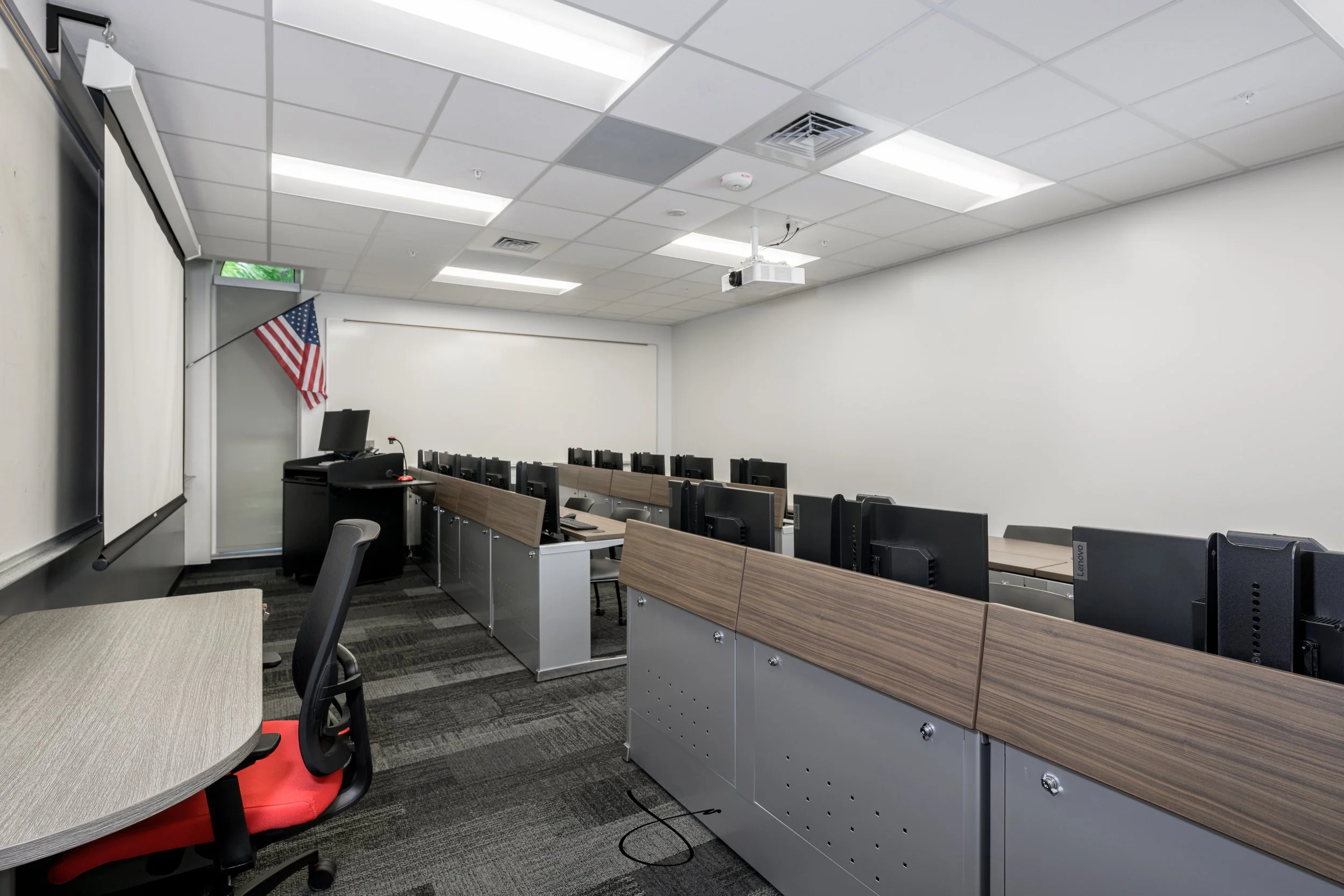 Classroom with rows of computers, a whiteboard, an American flag, and a ceiling-mounted projector.